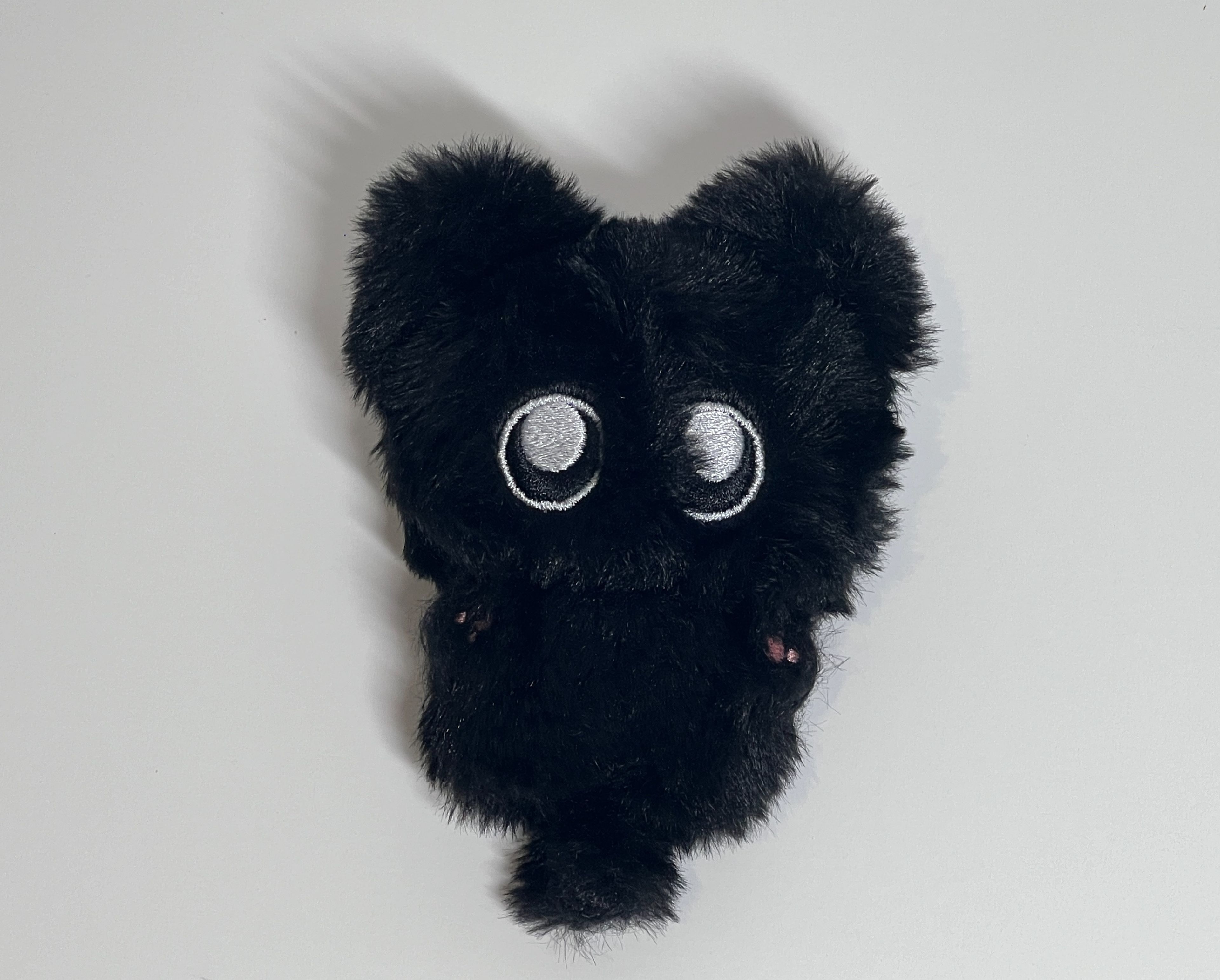 Close-up photograph of black plush character with embroidered white eyes against white background. Textural fur detail and minimal design emphasise the kawaii aesthetic of the soft toy.