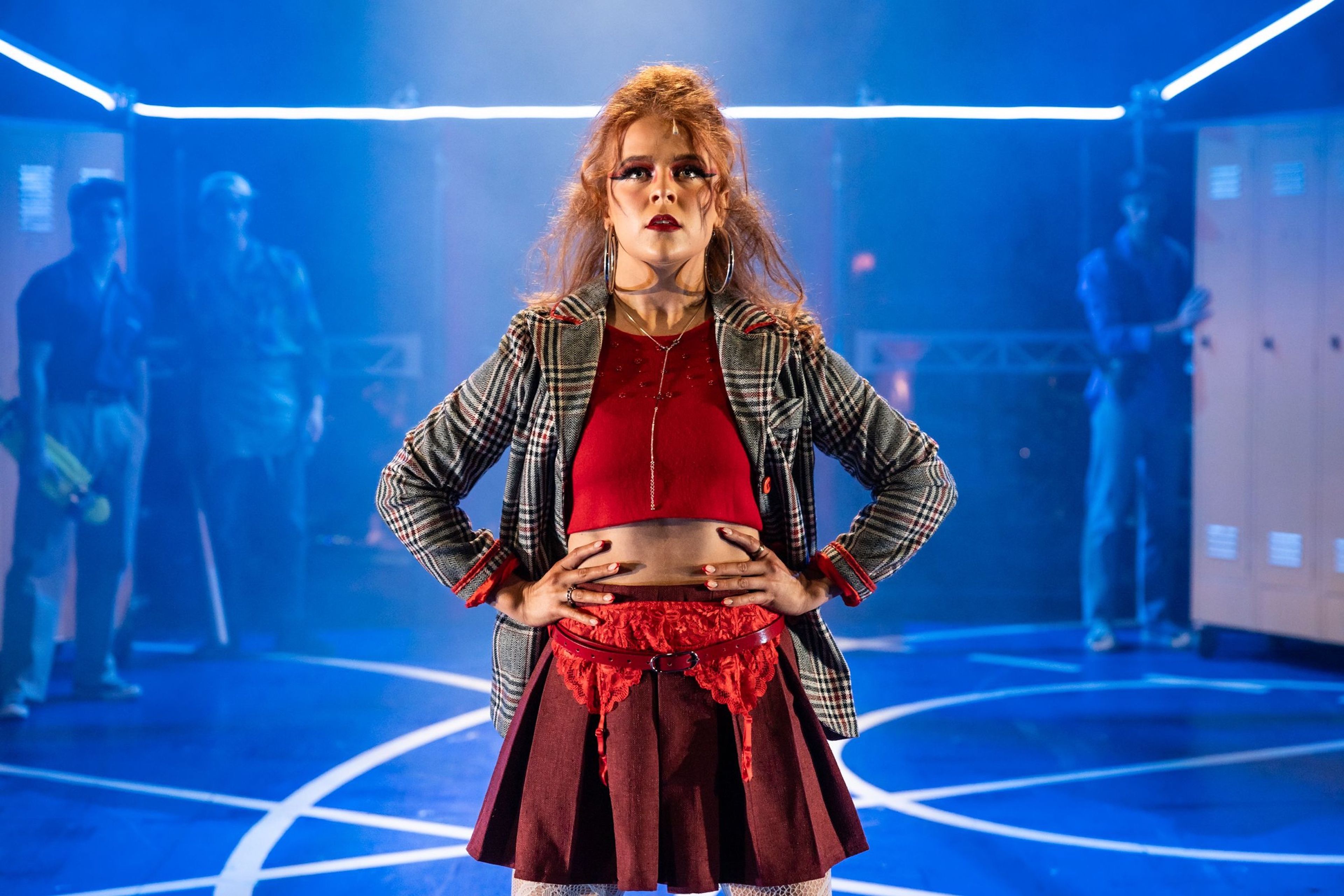 Theatre production photograph of a character in bold red and plaid attire standing confidently with hands on hips, set against a background with lockers and blue lighting.