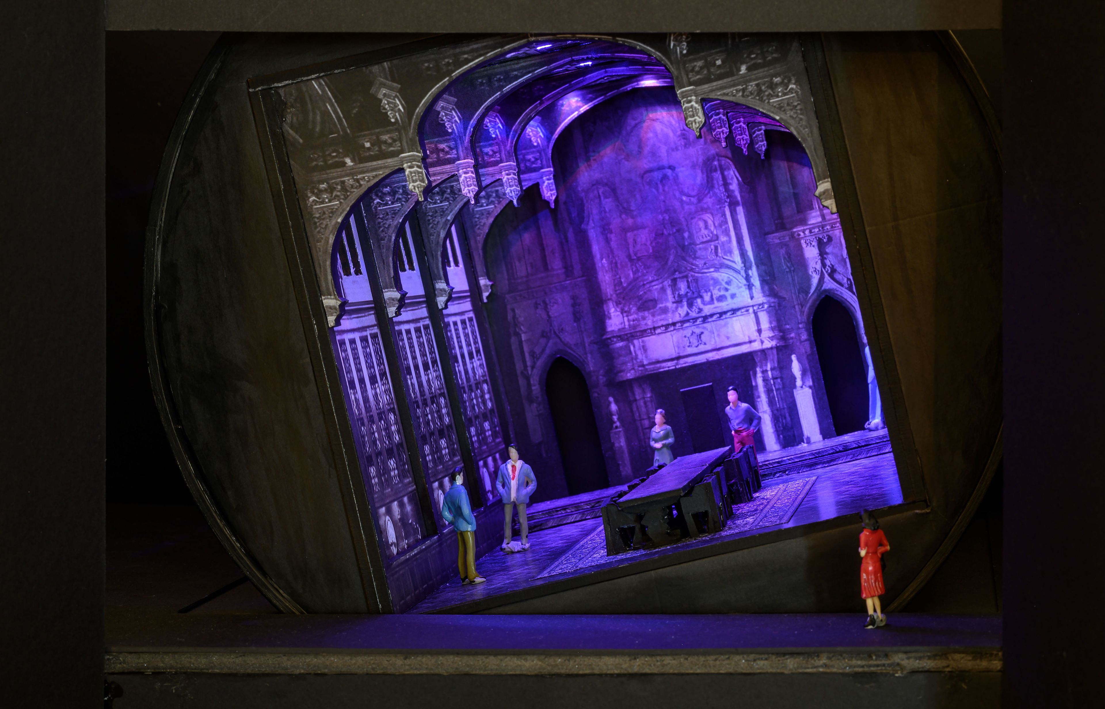 This image depicts an intricate stage model with a gothic architectural design, highlighted by ornate arches and detailed stonework. The set features a long table at the centre, surrounded by miniature figures representing characters in the scene