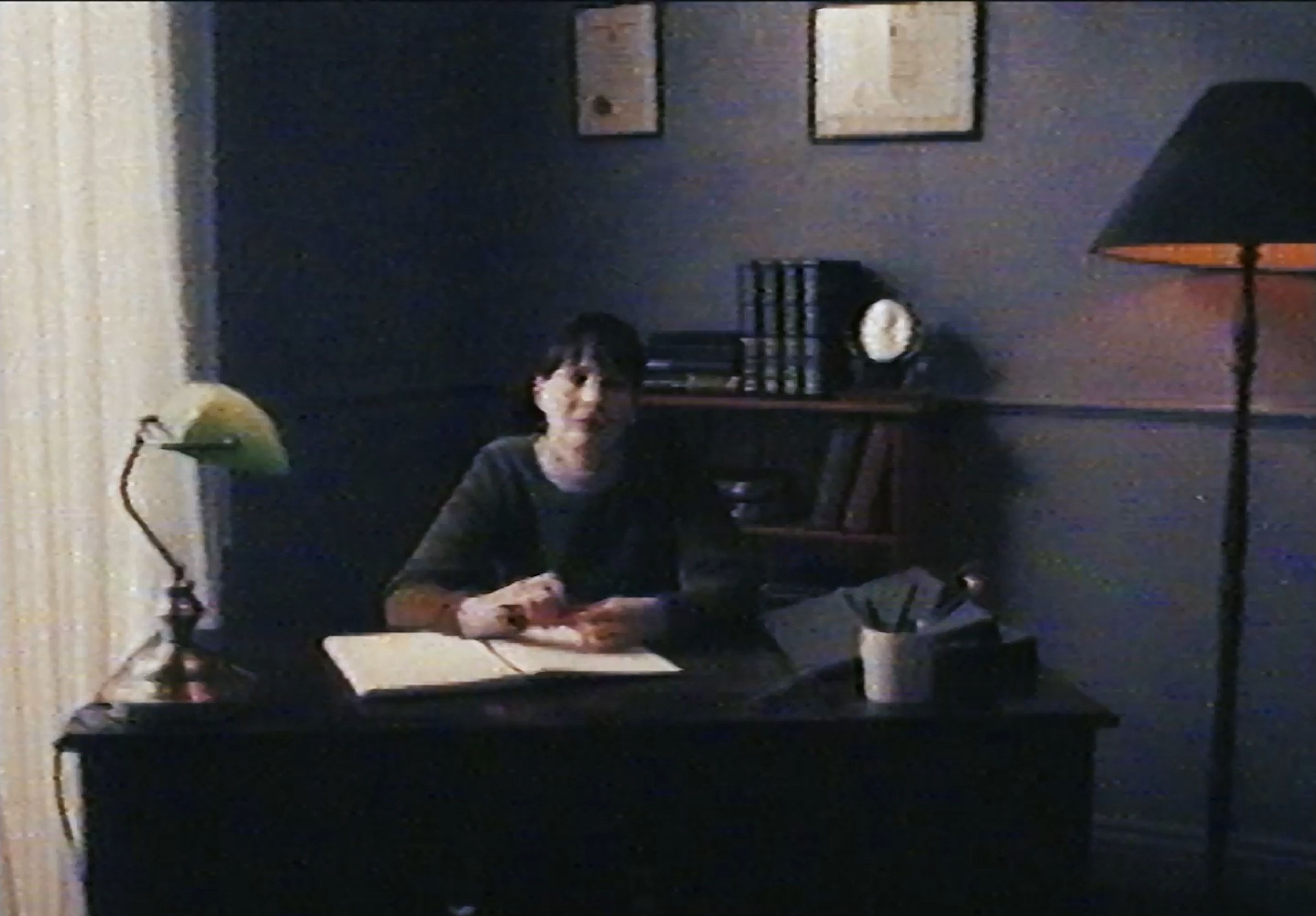 Film still shot on grainy vintage video showing a person at a study desk with banker's lamp and books in background.