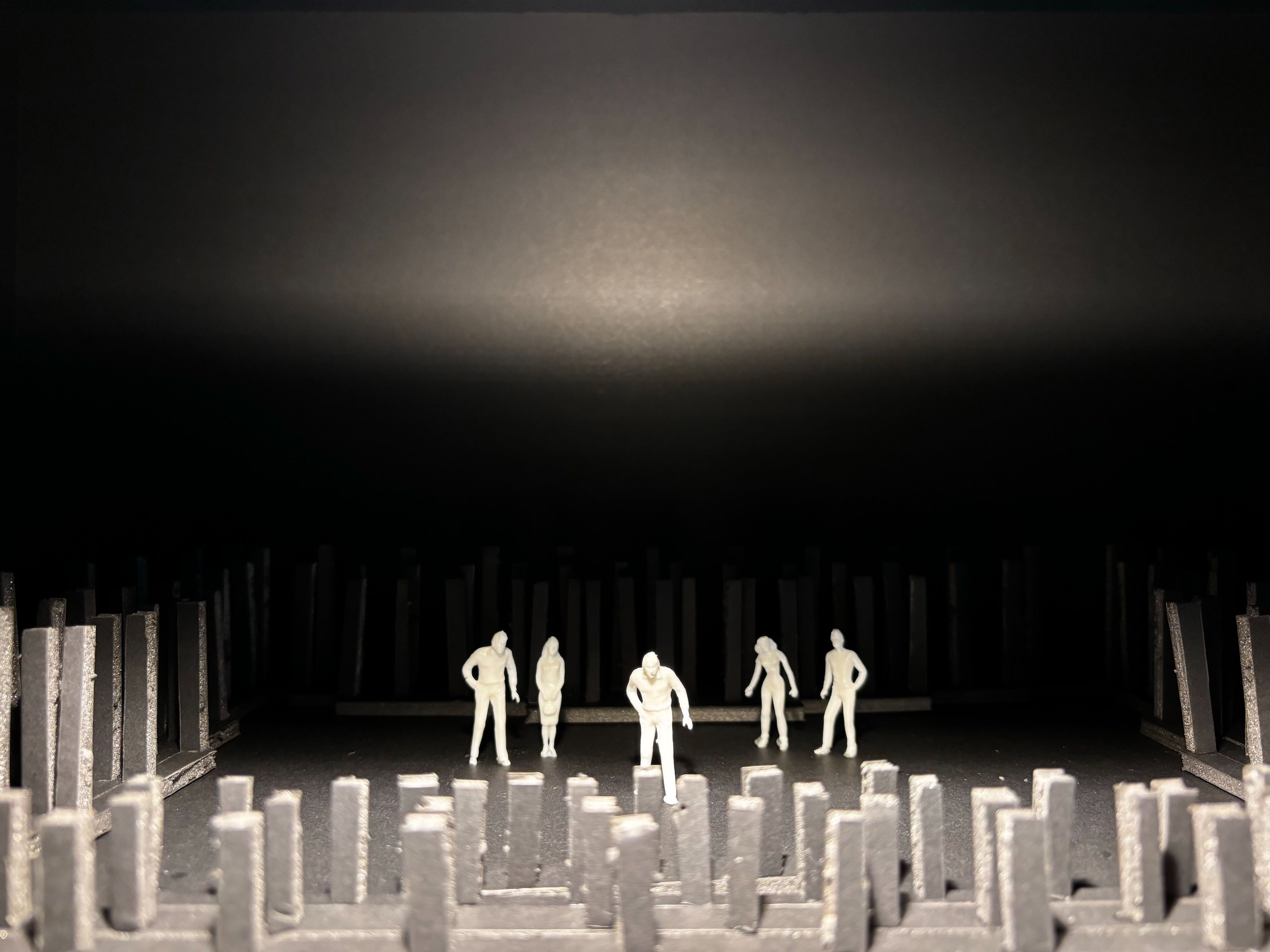 Scale model of a set design with white figures standing amongst vertical grey structures on a dark stage.