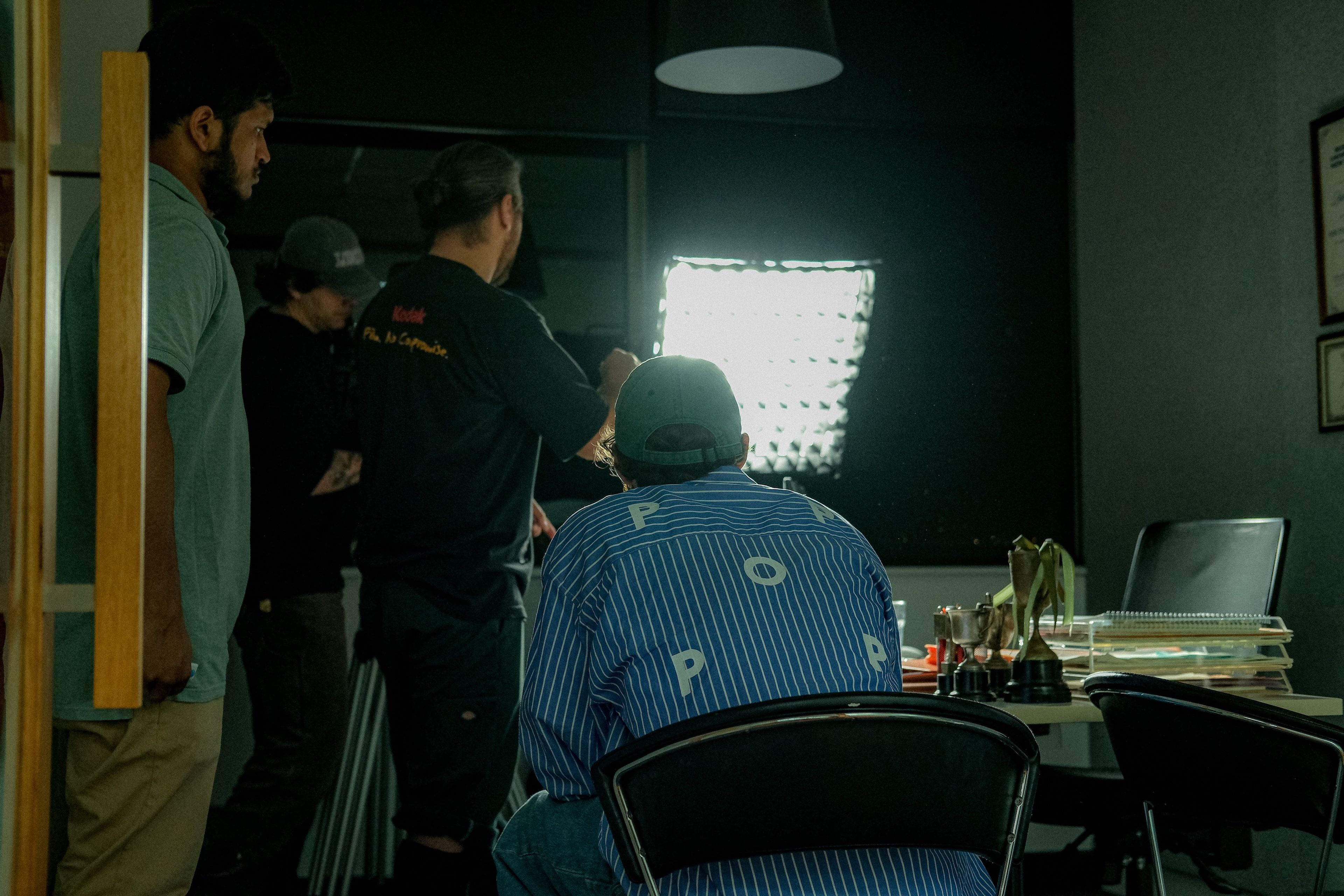 Behind-the-scenes still of a film crew setting up lighting equipment in a dimly lit room with a man seated in the foreground, wearing a striped shirt with "POP" text on the back.