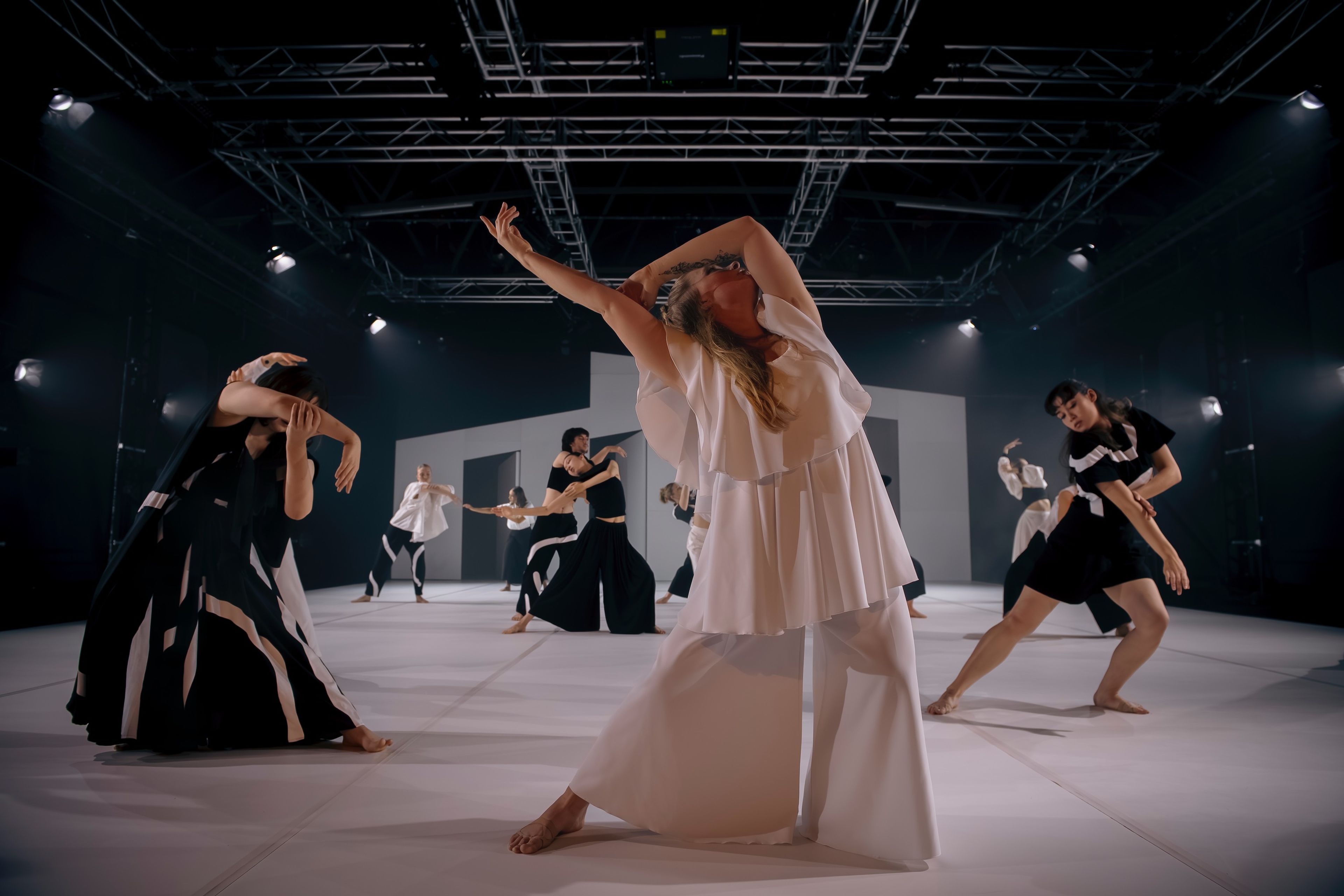 A contemporary dance performance with dancers in black and white costumes. The central dancer extends her arms dramatically, with other performers posed in expressive, angular positions.