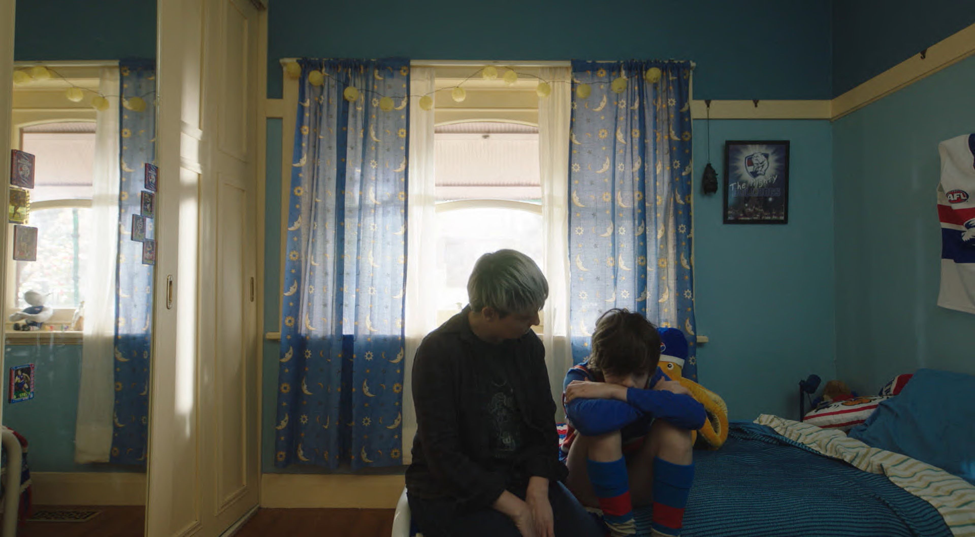 blue bedroom with patterned curtains featuring moons and stars. A young person sits on a bed with their head resting on their knees, while an older individual sits nearby offering comfort