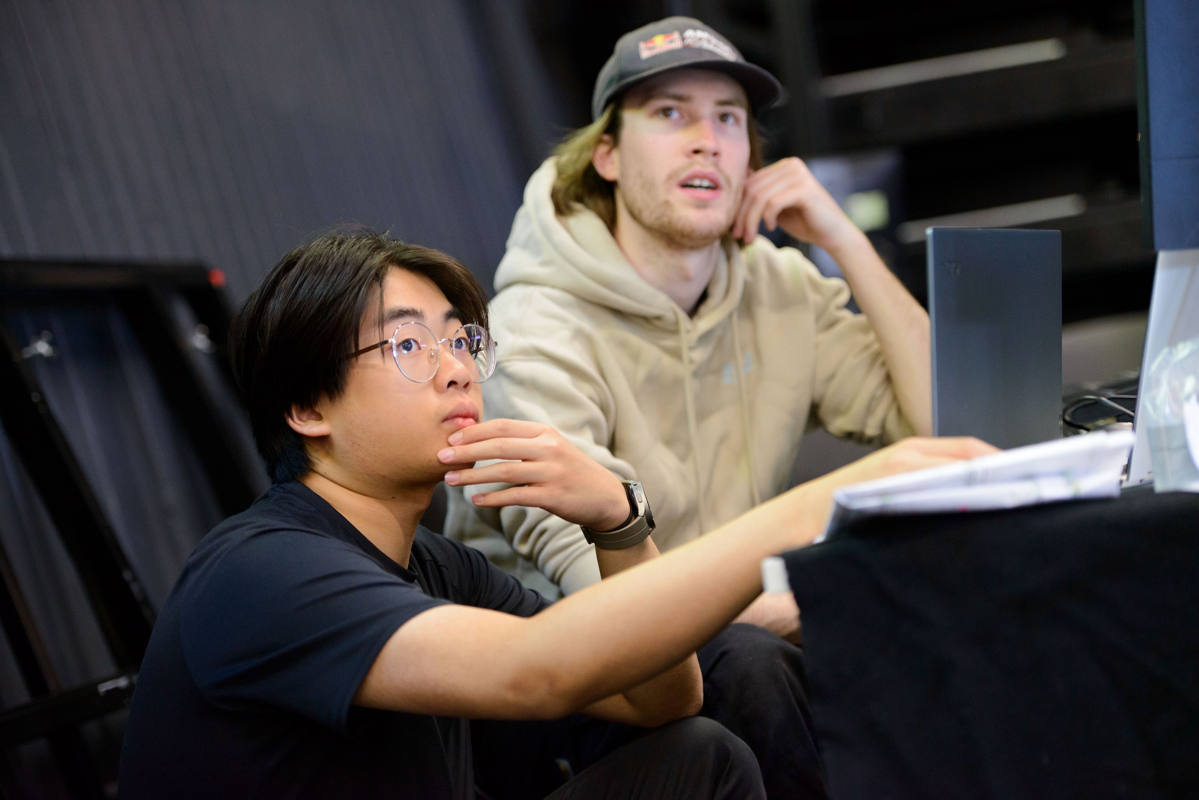 Close-up of two theatre crew members looking attentively at a screen or equipment off-camera. One wears glasses and a black shirt, the other a cap and beige hoodie, both appearing focused and engaged in their task.