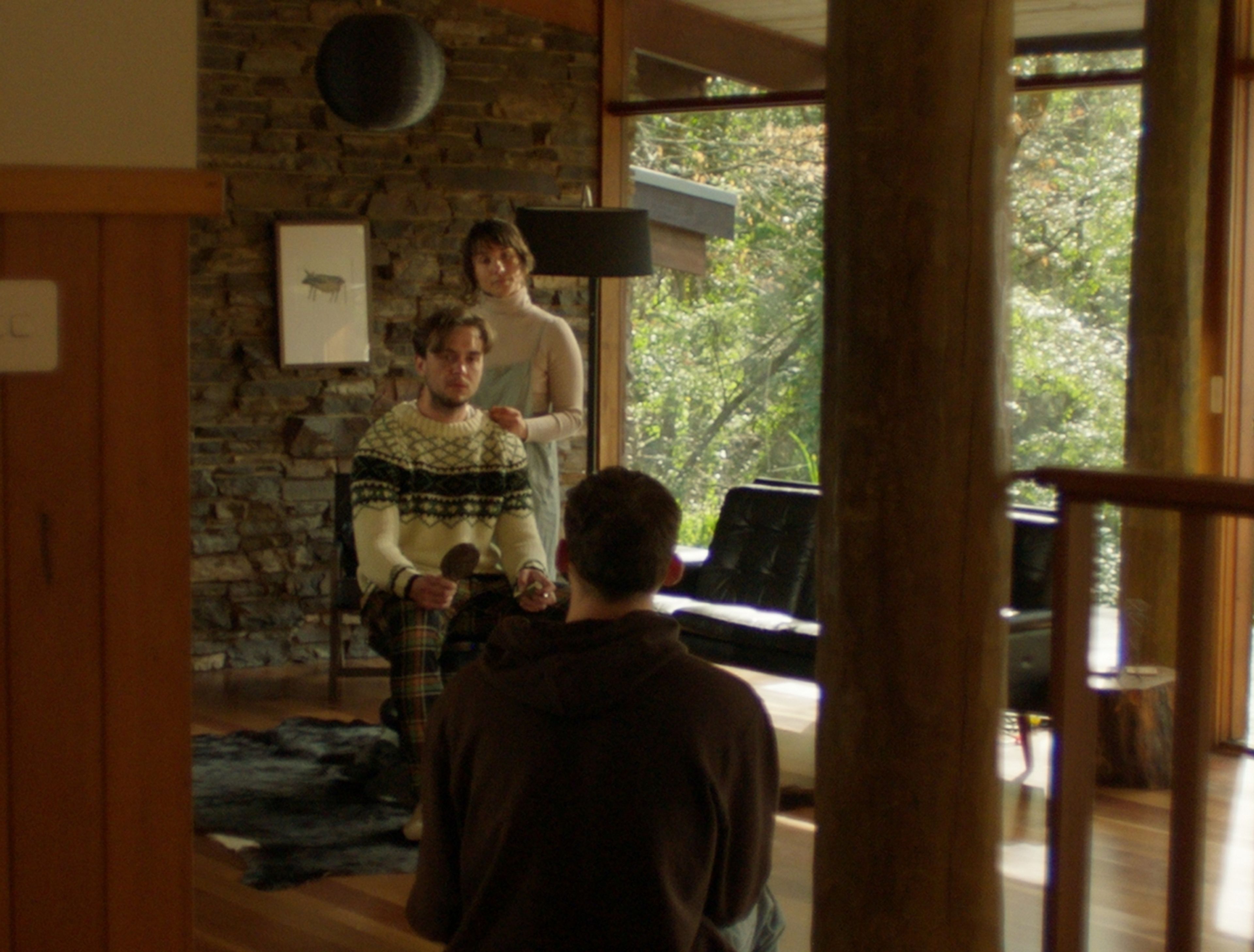 Film still of a modern cabin interior featuring stone walls and floor-to-ceiling windows, with three people in winter knitwear near a grand piano, bathed in golden afternoon light.