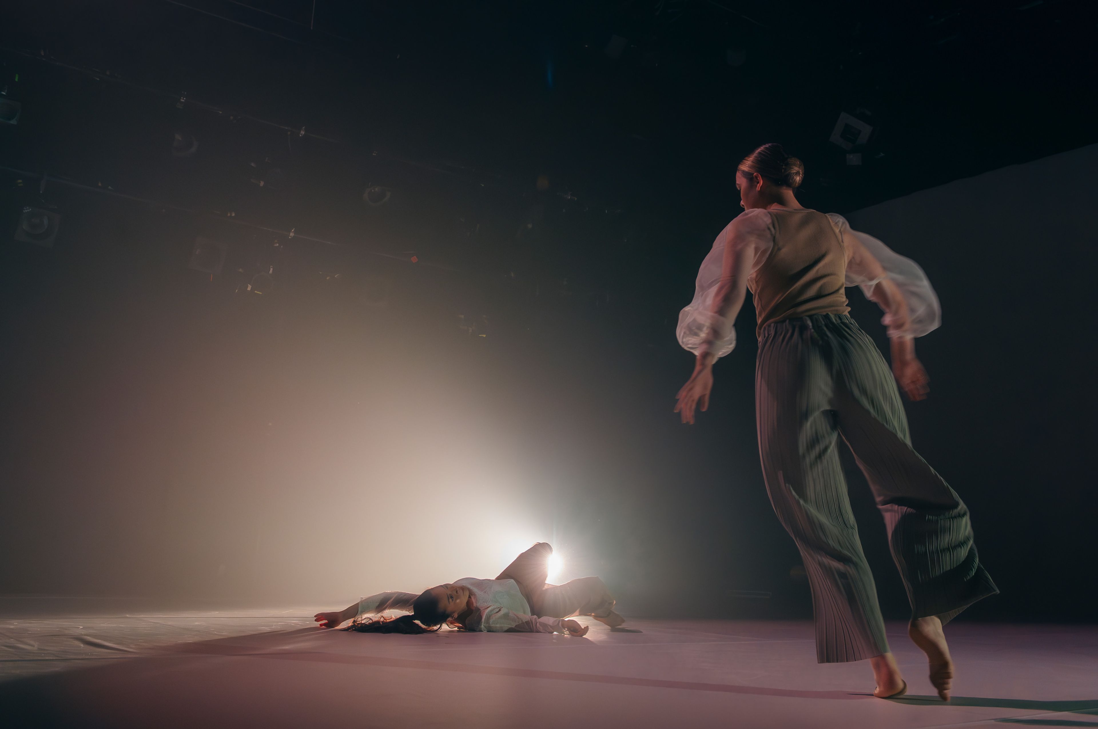 Dance production photograph featuring a dancer lying on the floor with another dancer approaching, light casting a gentle glow behind them.