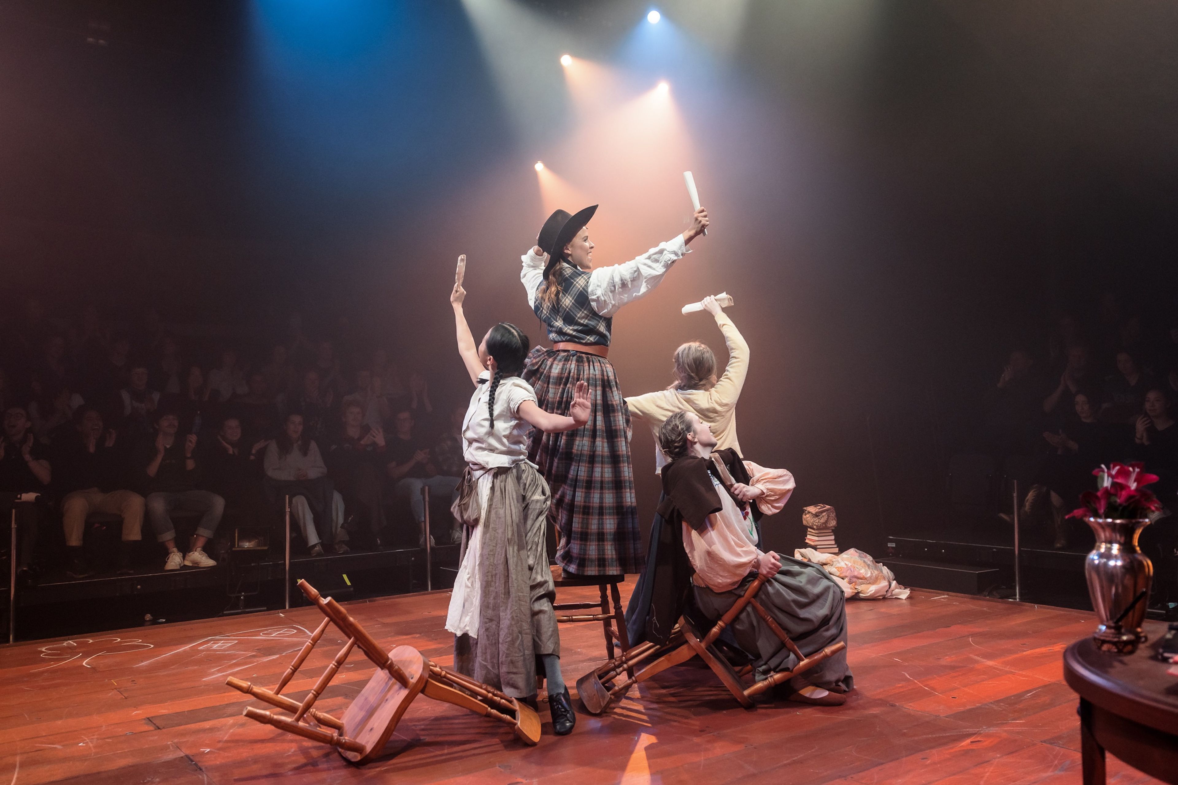 Theatre production photograph showing four actors in period costumes posing triumphantly, holding objects aloft, with a warmly lit audience in the background applauding.