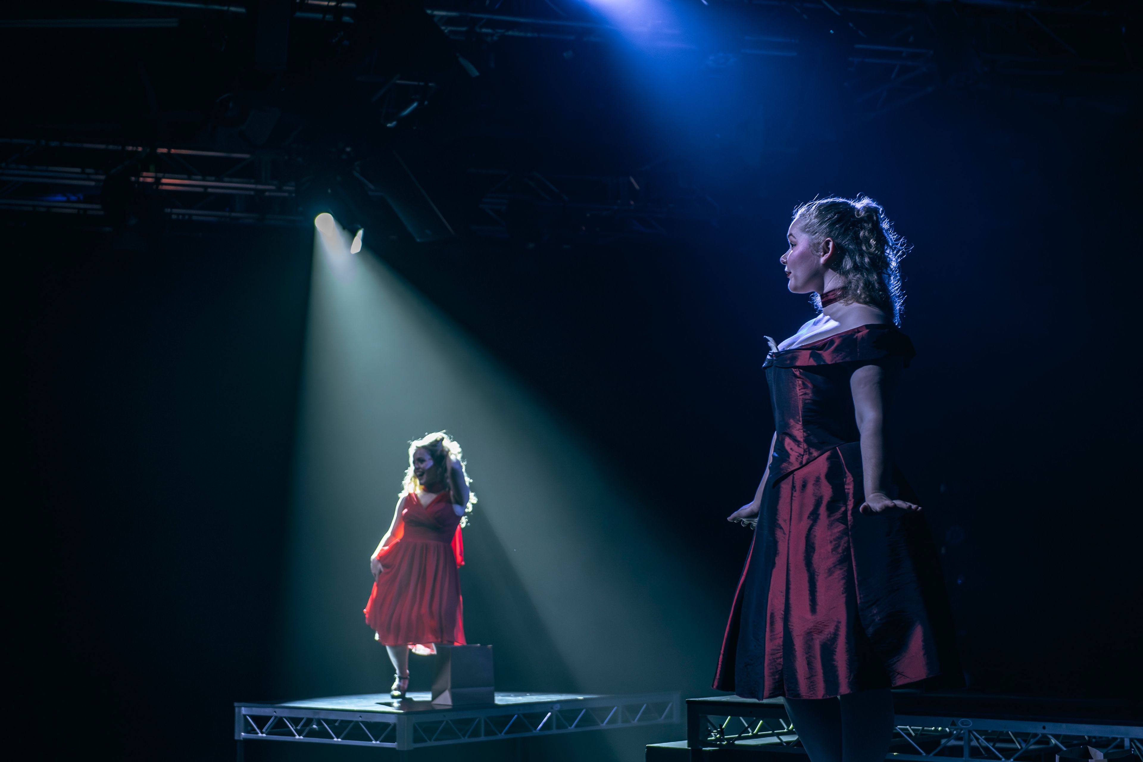 Theatre production photograph showing two actors in red dresses standing on separate platforms, with a dramatic spotlight illuminating one actor from above.