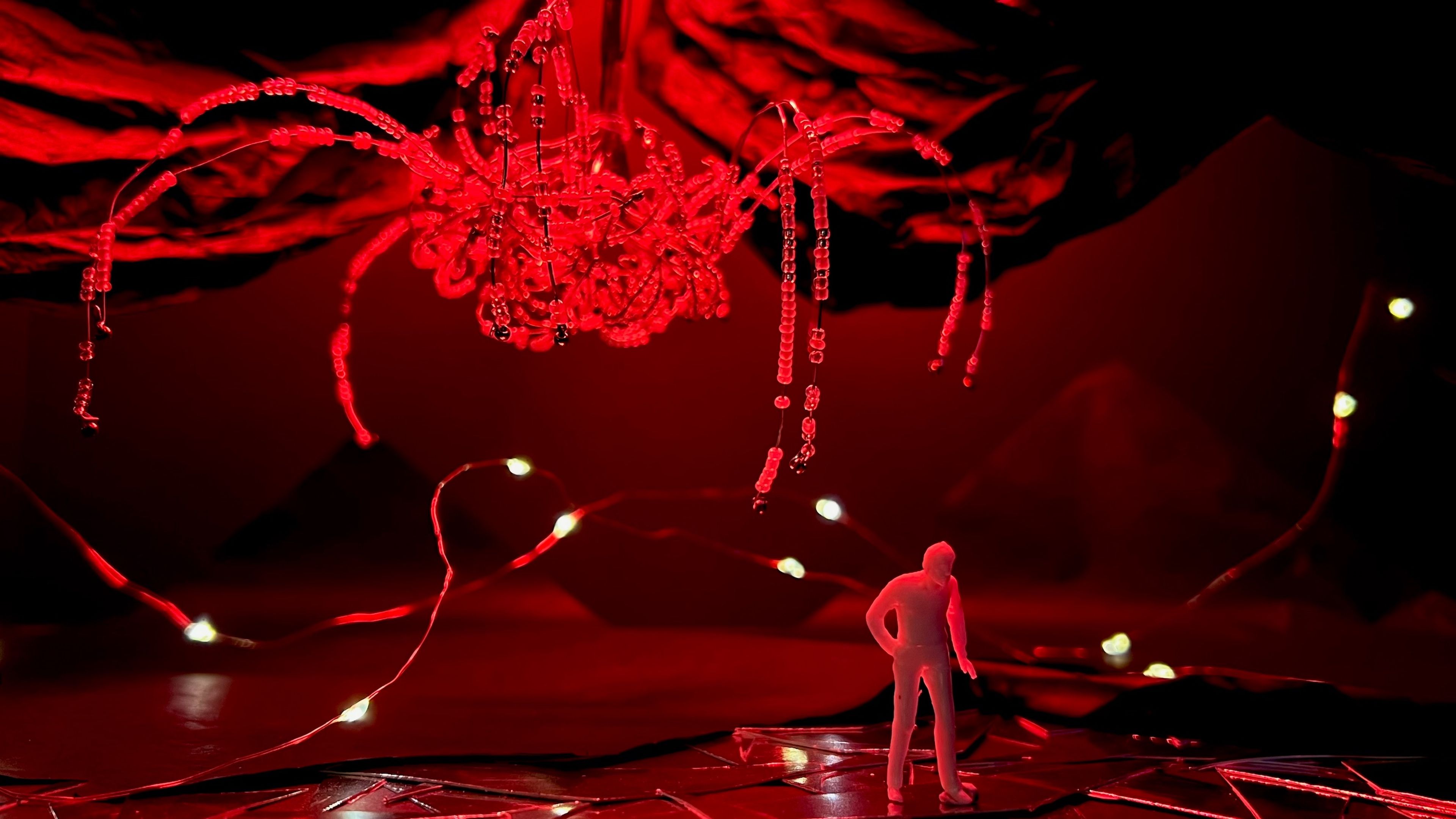 Scale model of a theatrical set with a red figure standing beneath a suspended chandelier-like structure made of beads, illuminated in dramatic red light against a dark background