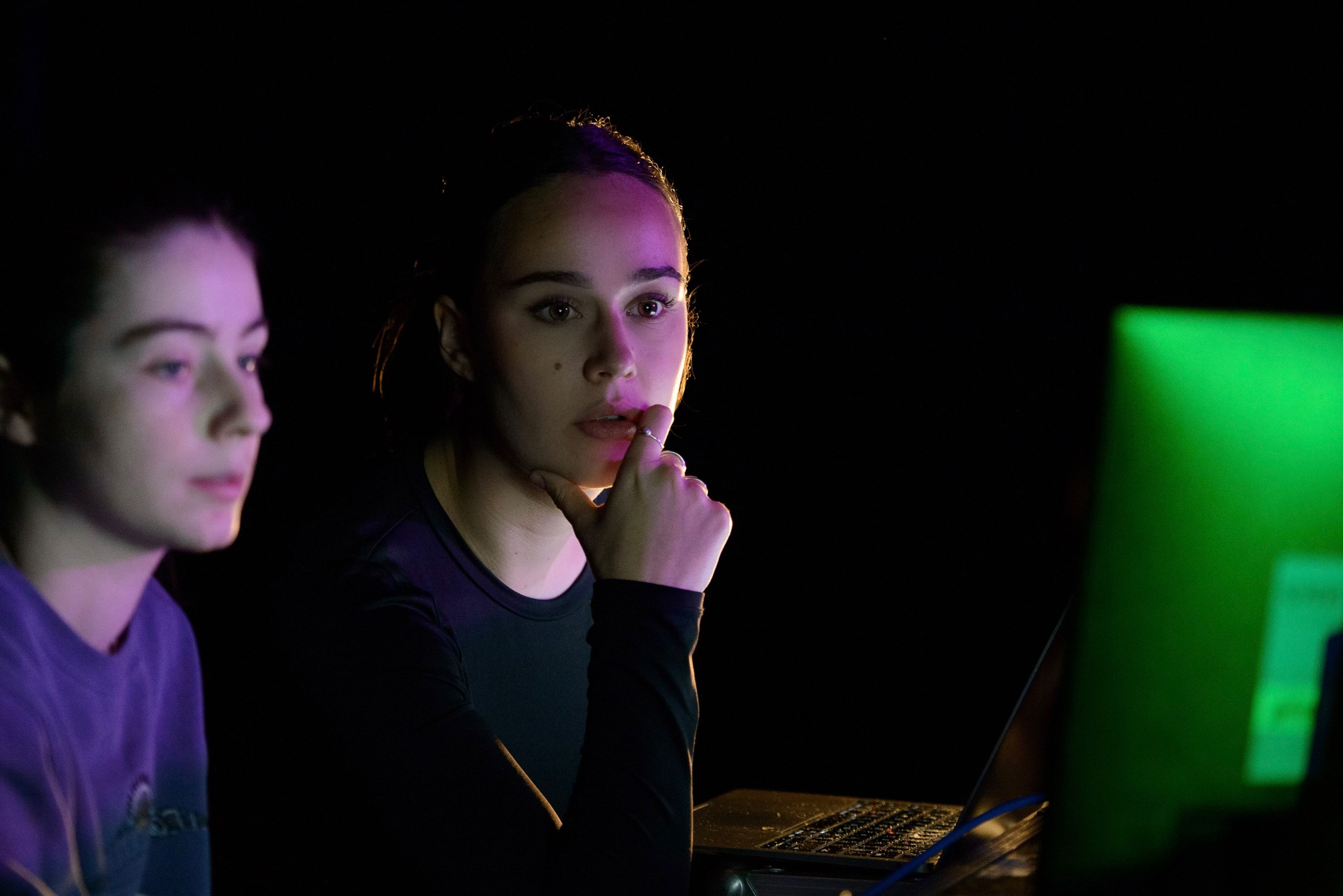 Two people sit in dim light, one thoughtfully resting a hand on their chin, illuminated by purple and green light, with a laptop visible nearby