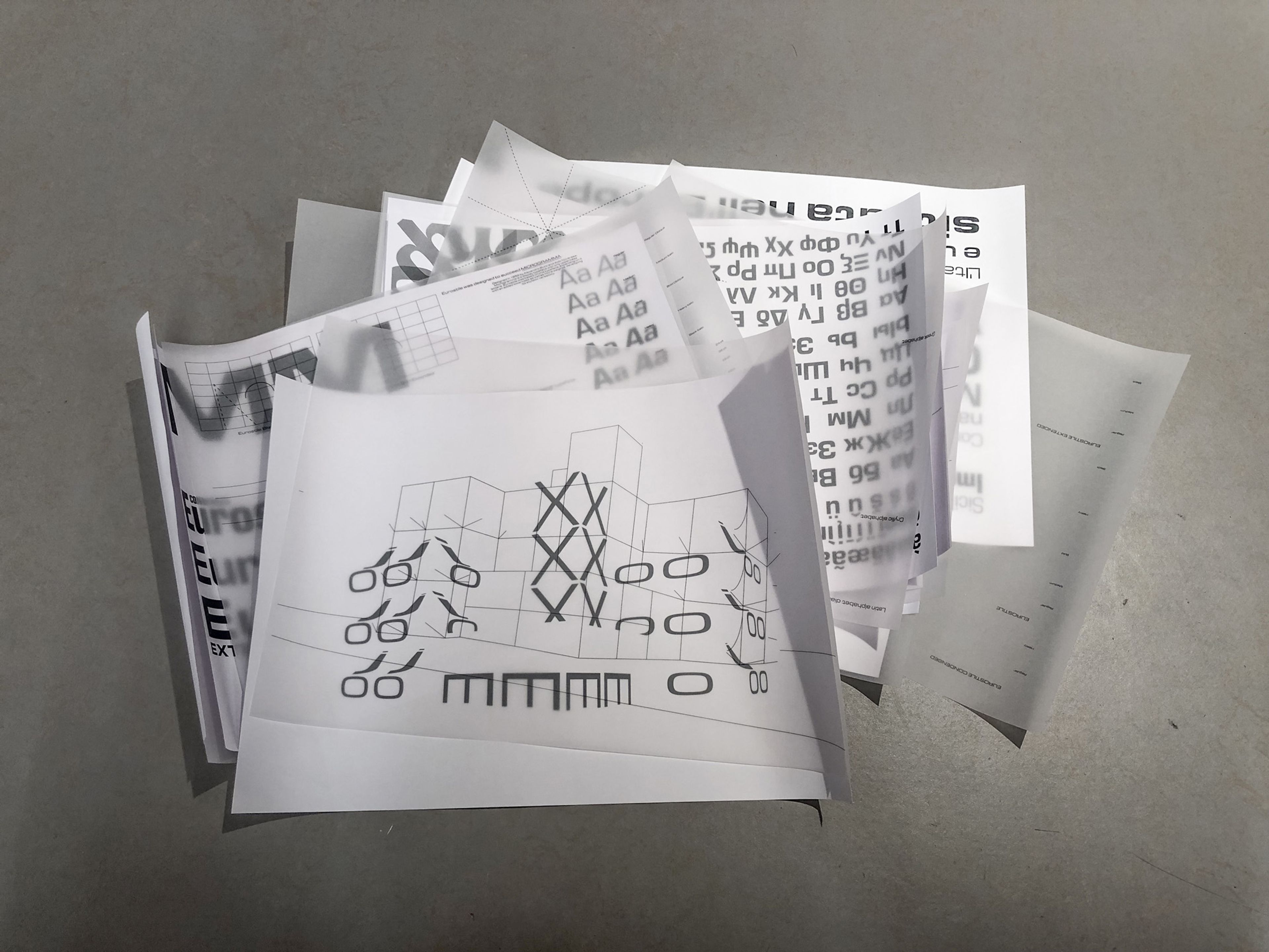 A scattered pile of transparent and printed sheets featuring various typographic designs, grids, and text layouts, placed on a neutral grey surface