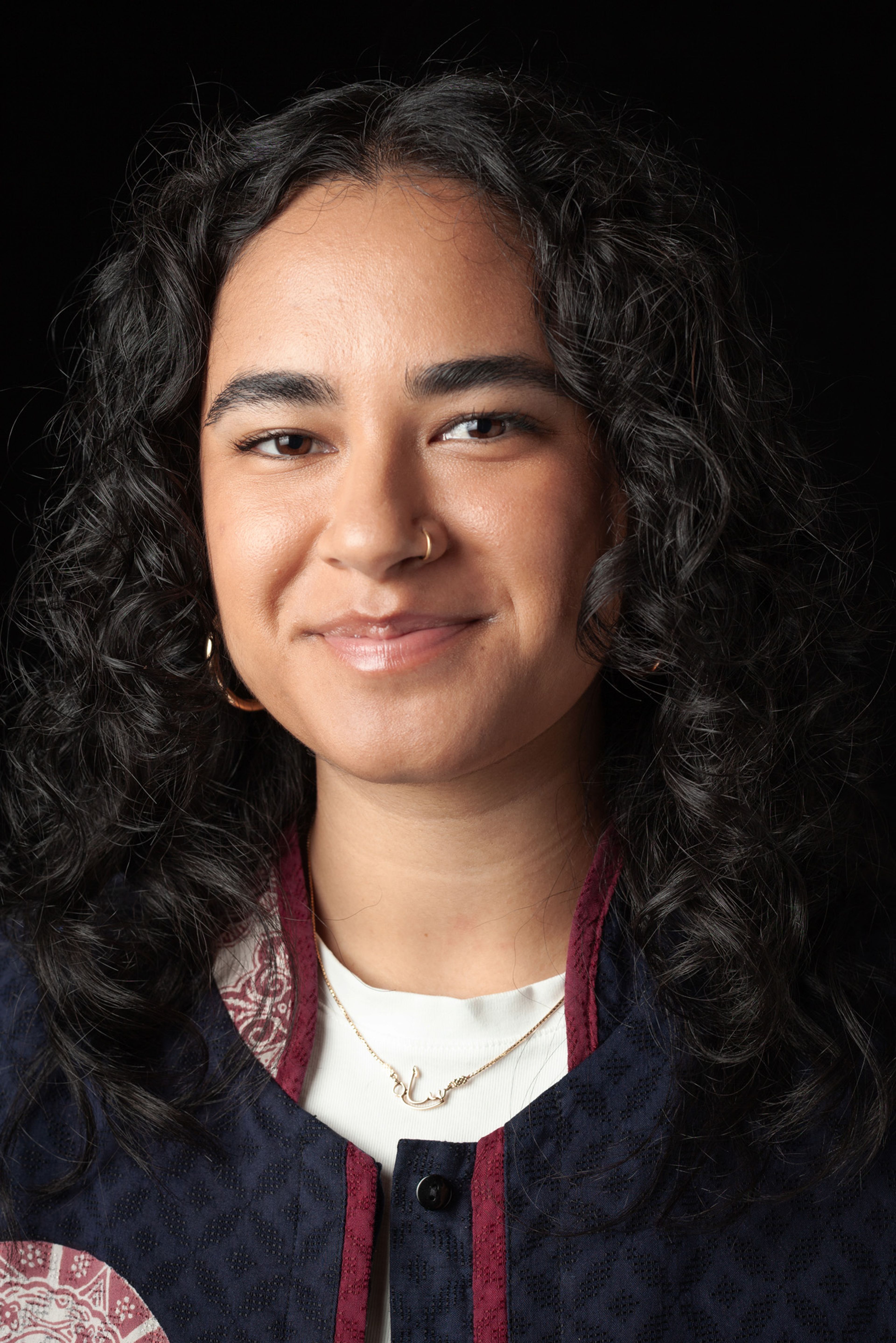 Headshot of a young woman with curly black hair against a dark background, wearing a navy and burgundy patterned jacket, a white top, and a delicate gold necklace.