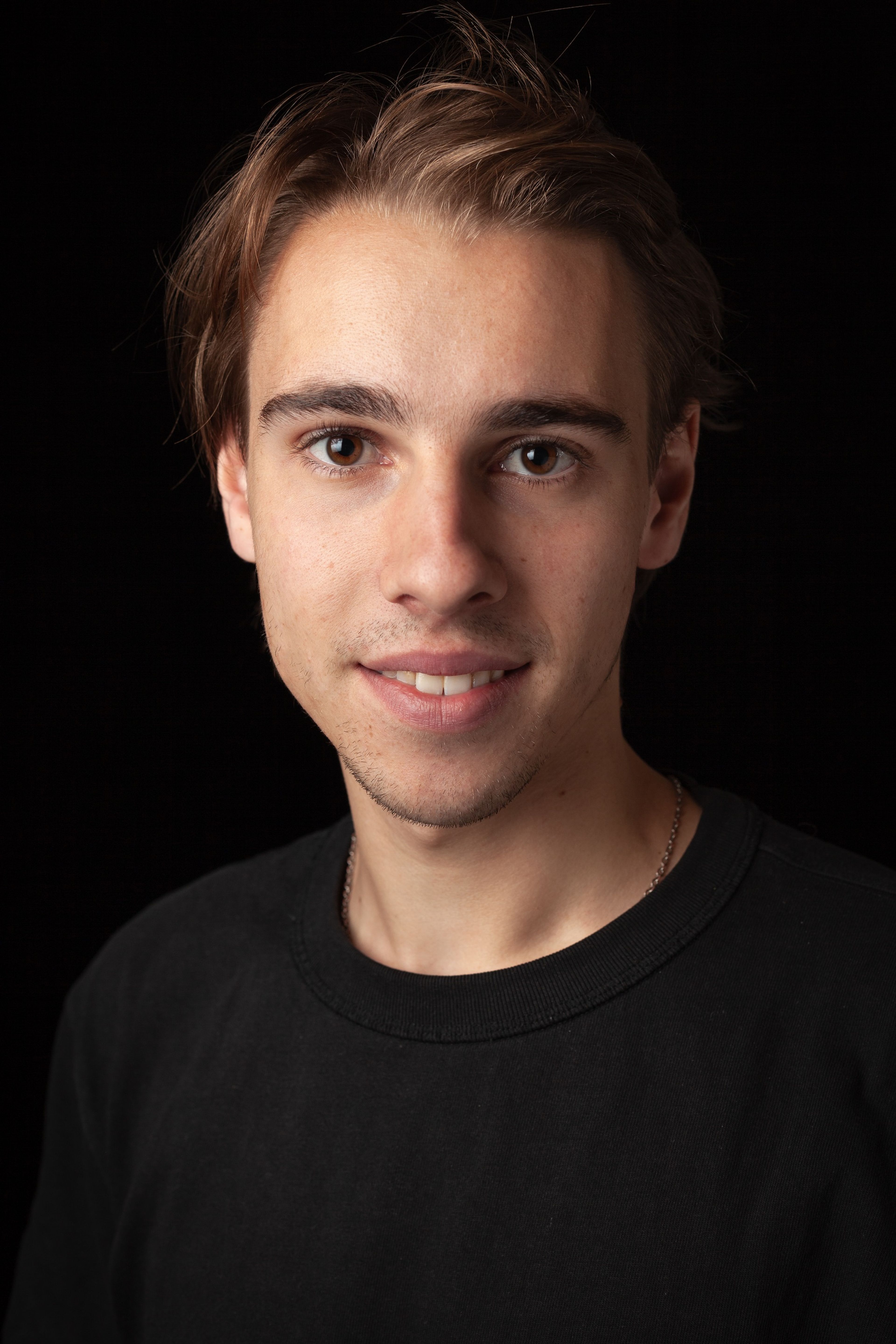 Headshot of a young man with short light brown hair, wearing a plain black shirt, against a black background.