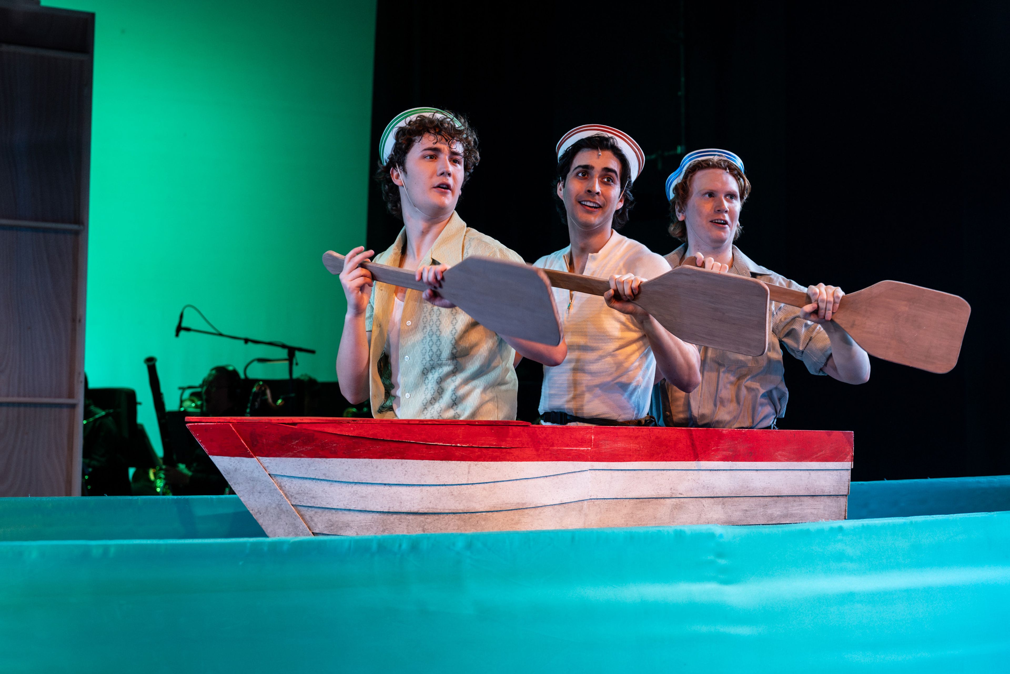 Theatre production photograph showing three performers in sailors' caps rowing a stylised red and white boat prop against a green-lit backdrop.