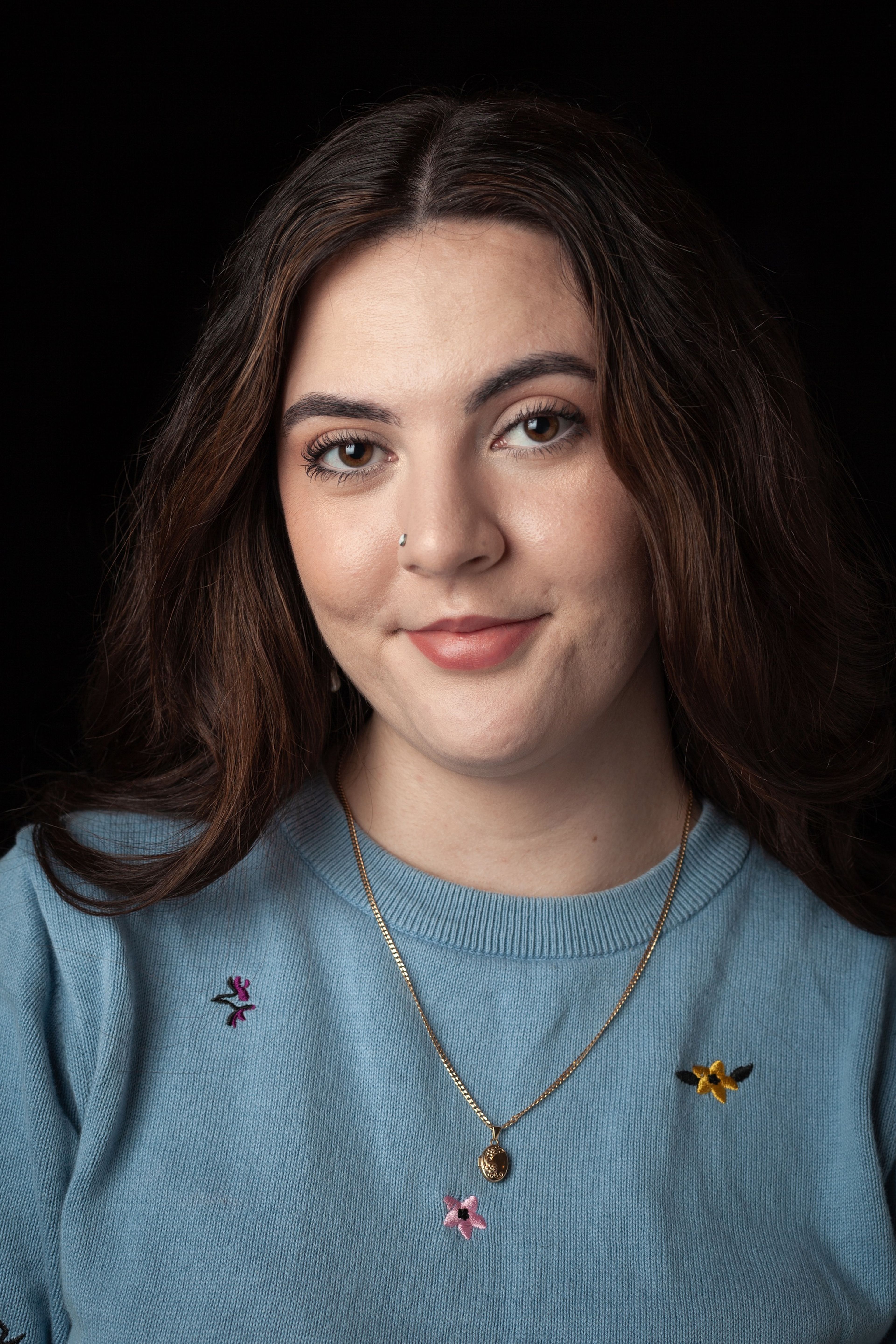 Headshot of a young woman with long dark hair, wearing a blue jumper with small embroidered flowers and a gold necklace, against a black background.