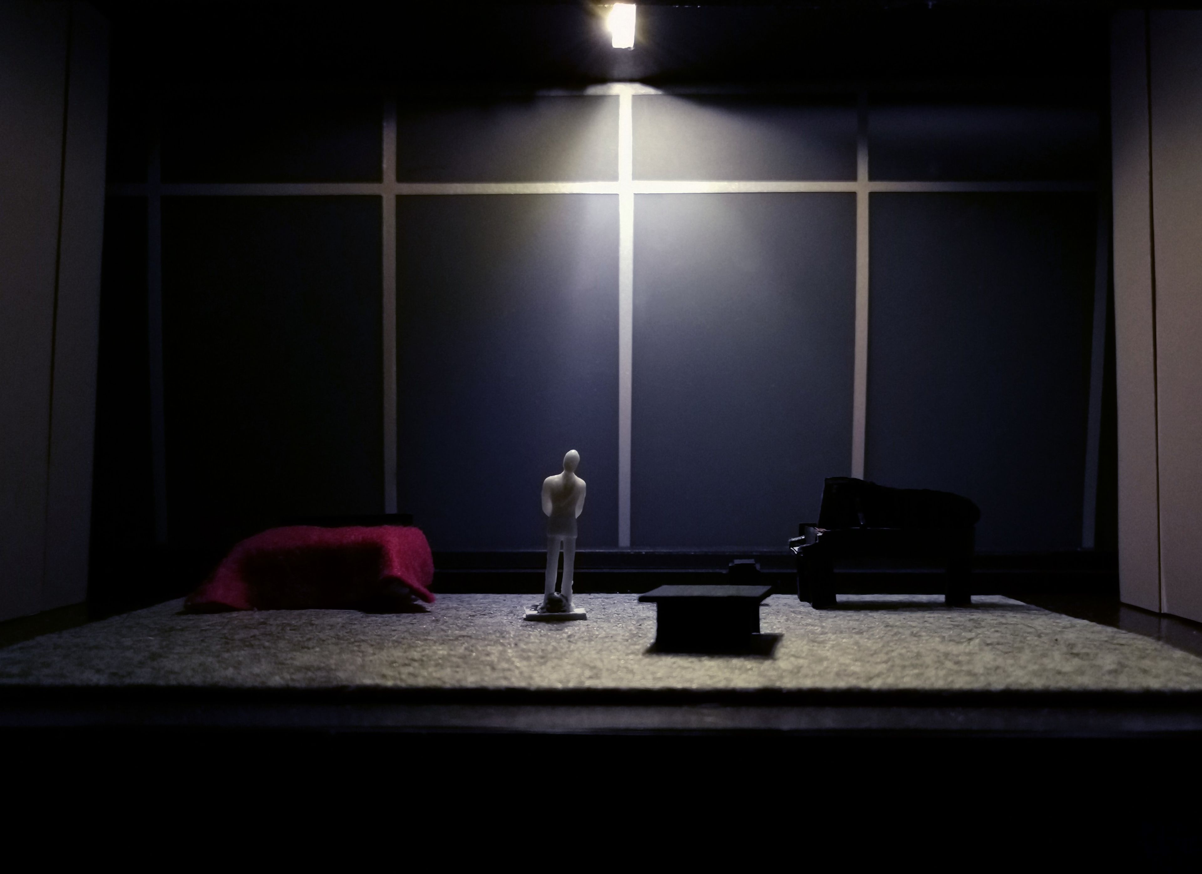 Scale model of a set design illuminated by moody, atmospheric lighting, highlighting a figure in the centre surrounded by dark furniture against a grey backdrop.