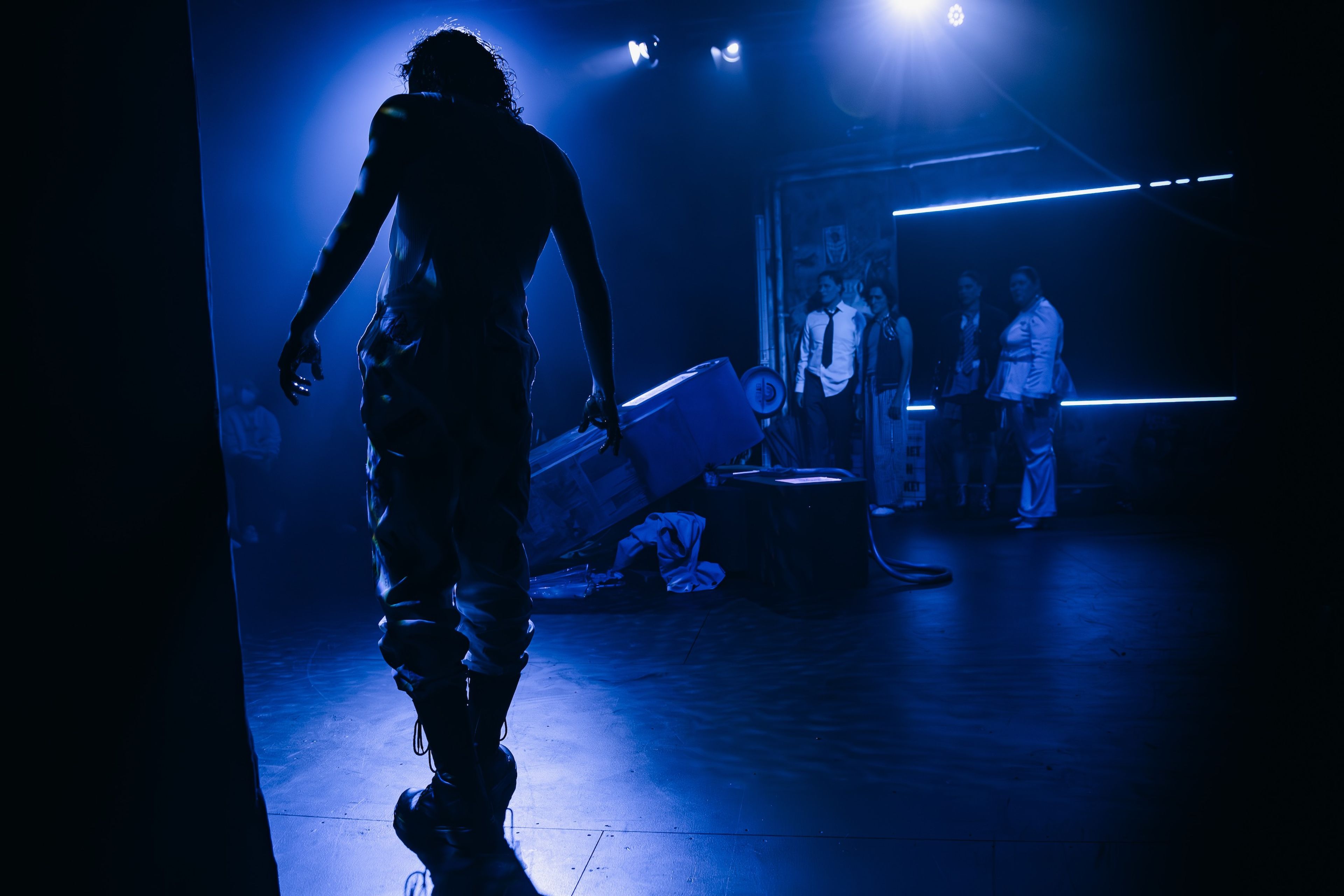 Theatre production photograph of a silhouetted male figure in a dark, blue-lit scene, with onlookers in the background.