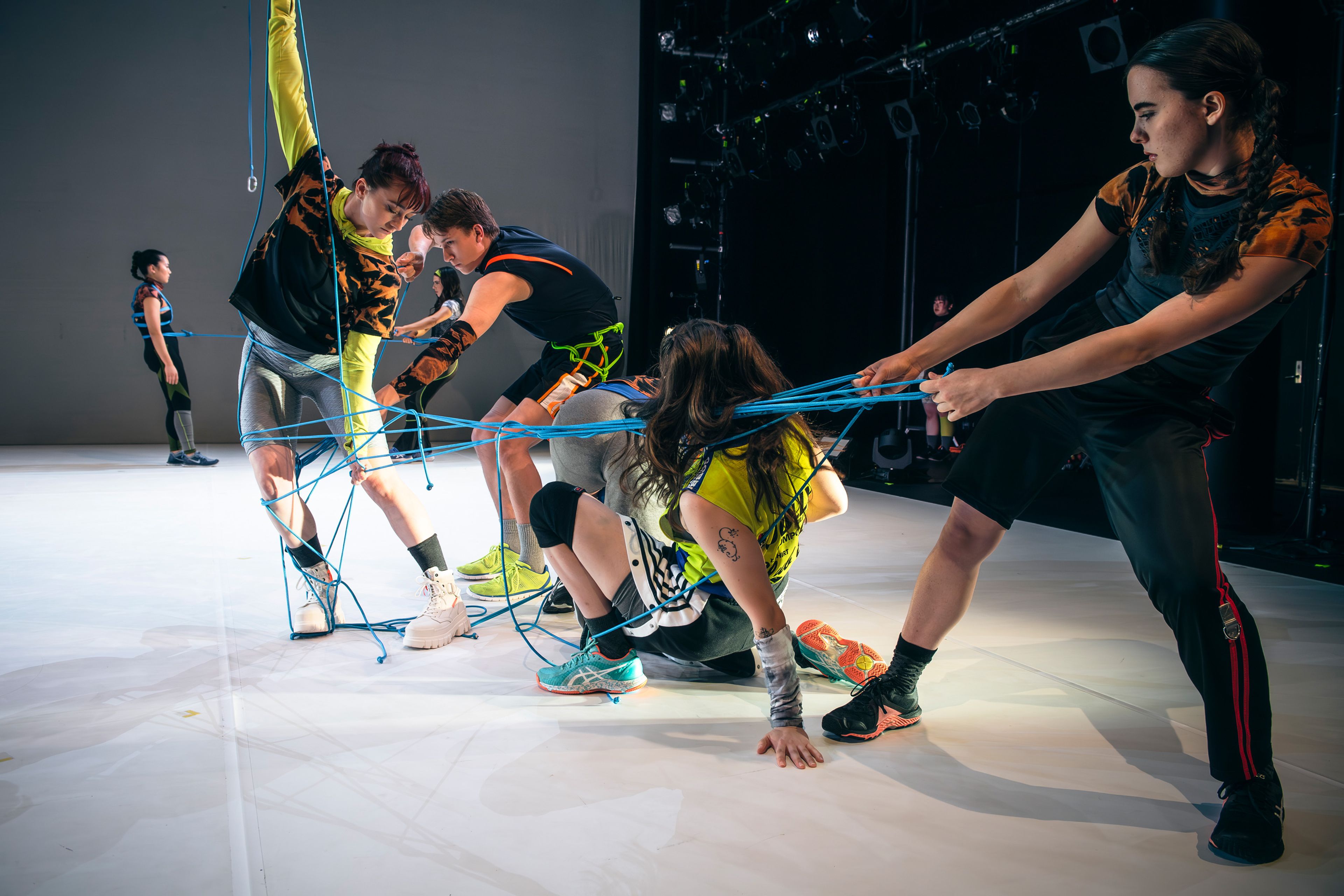 Dance production photograph showing a group of dancers entangled in blue ropes, pulling in different directions with focused expressions.