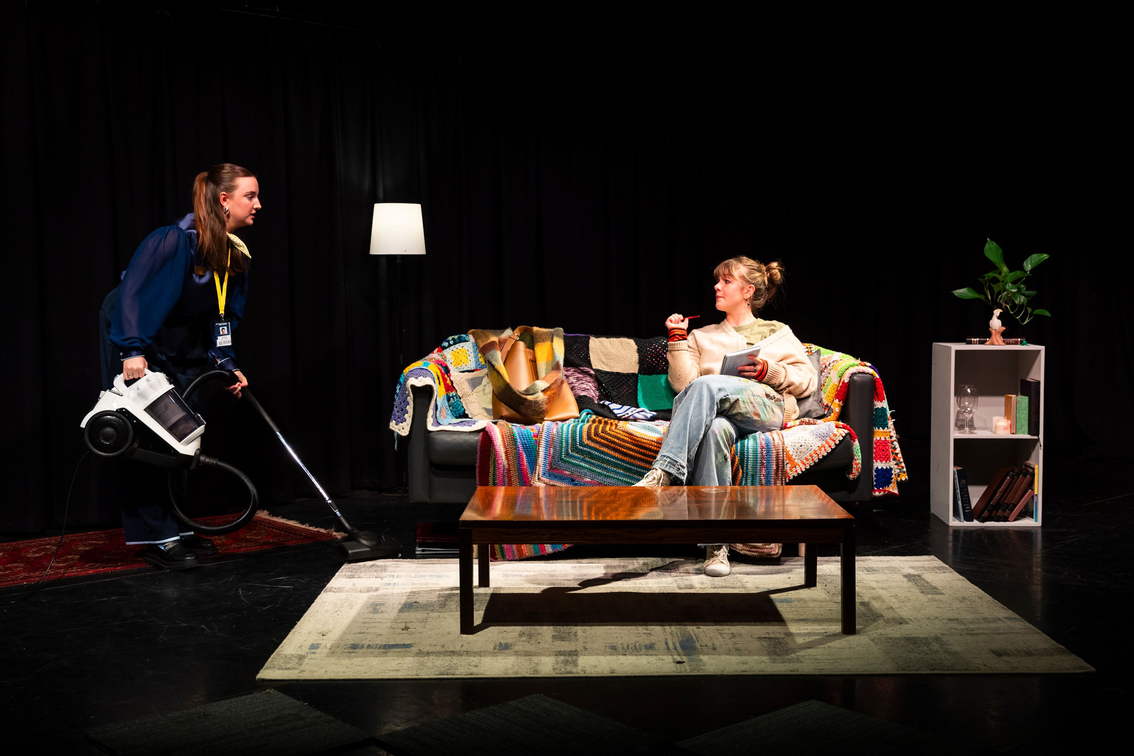 Theatre production photograph showing a domestic scene with a performer in casual clothes lounging on a couch covered in colourful quilts, while another performer in corporate wear operates a vacuum cleaner.