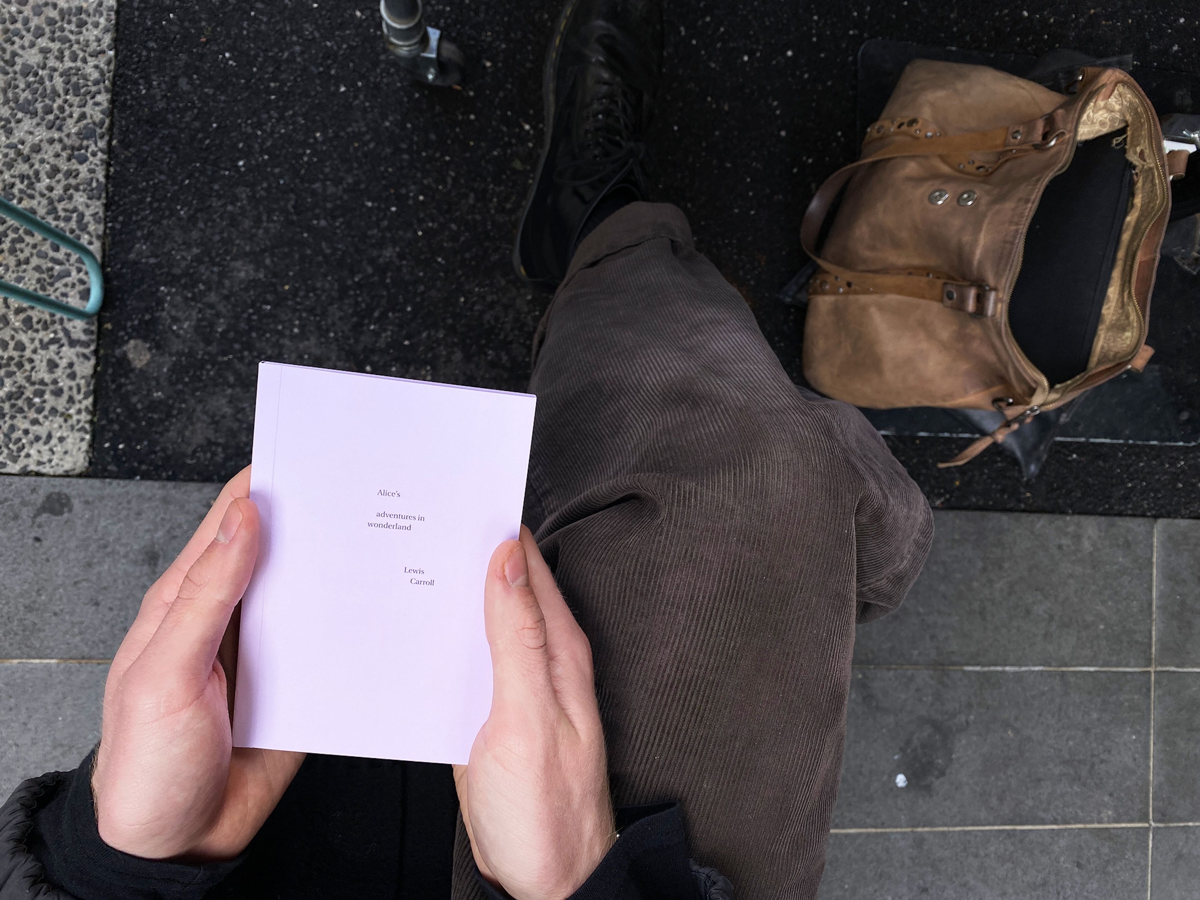 Close-up of a person holding a copy of "Alice’s Adventures in Wonderland" with a minimalist cover, seated outside.