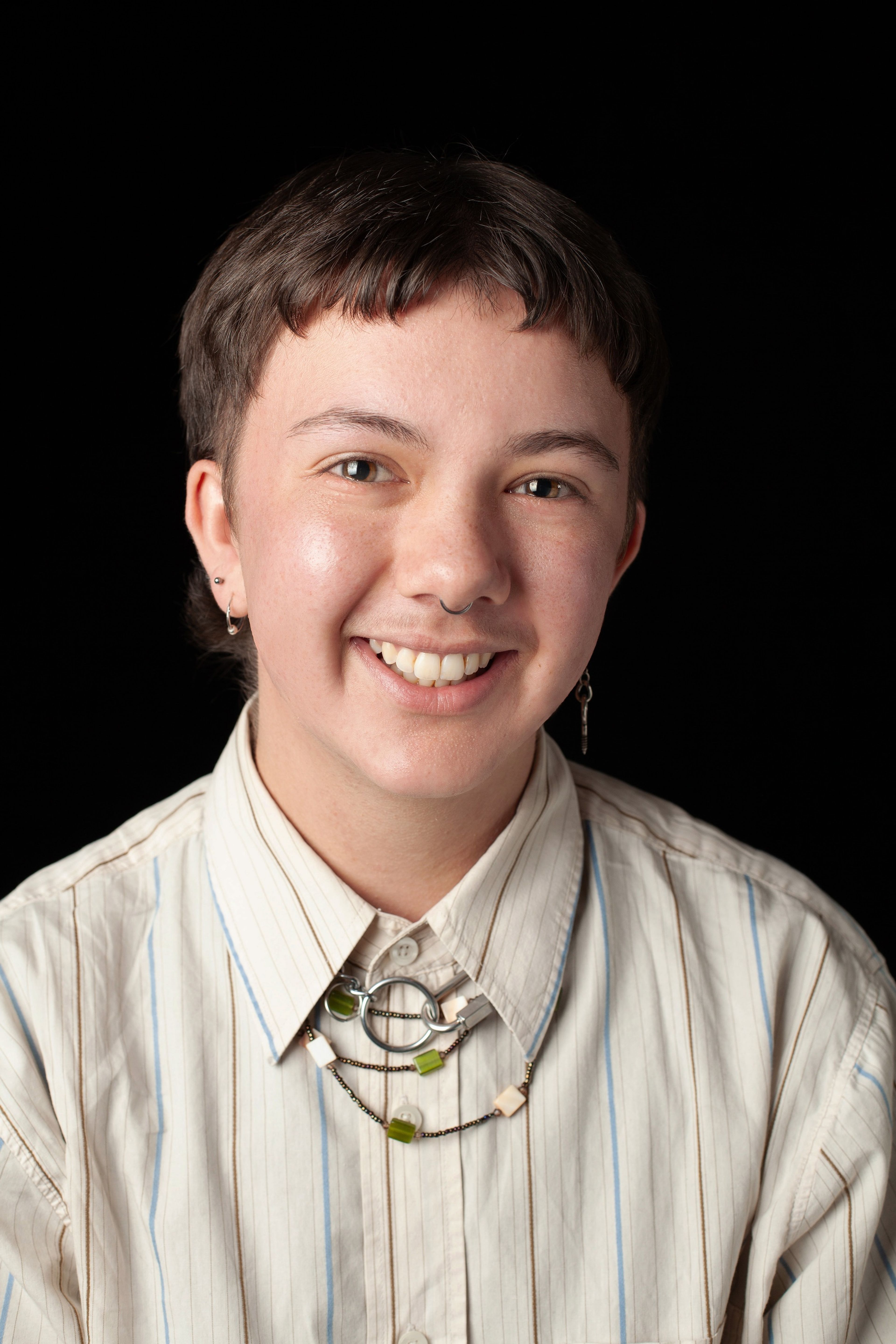 Headshot of Charlie. They smiling and have short dark hair, and are wearing a striped button-up shirt with a layered necklace, against a black background.