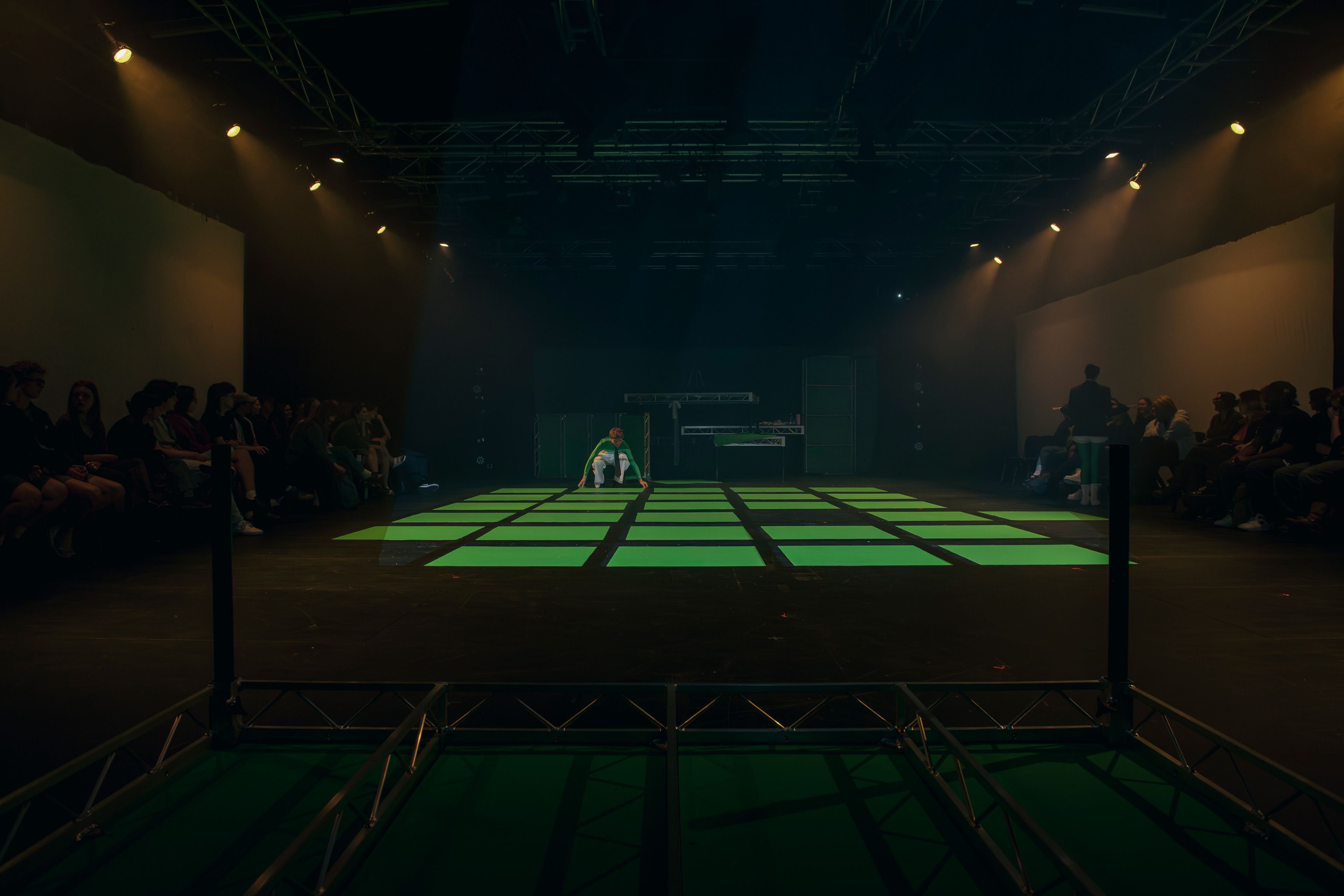 A dimly lit stage featuring a grid of glowing green squares as the central focal point. A performer crouches at the far end of the grid, while the audience is seated on either side in shadowy outlines.