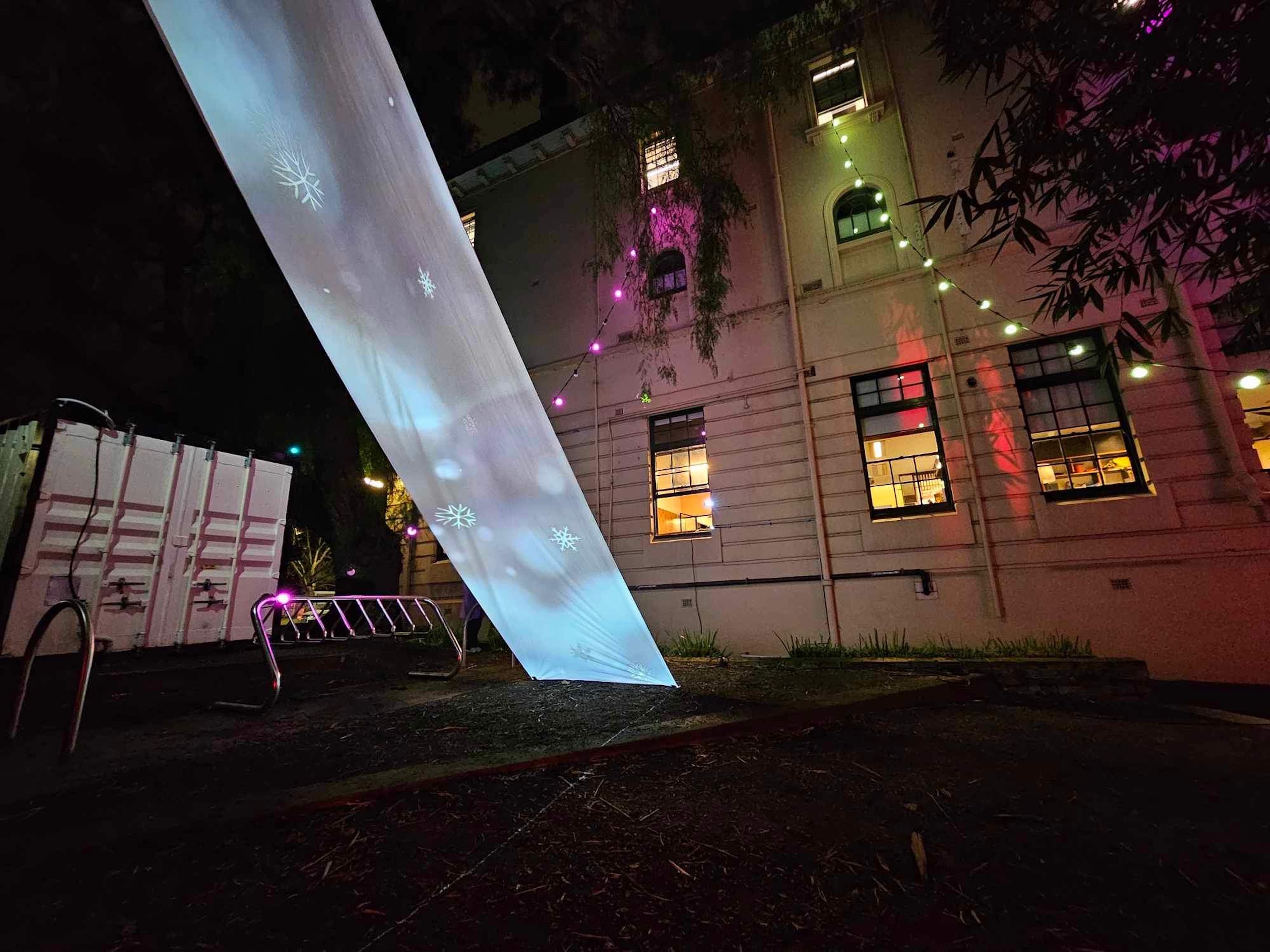 Outdoor installation with a large screen projecting snowflakes, and colourful lights strung against a building at night.