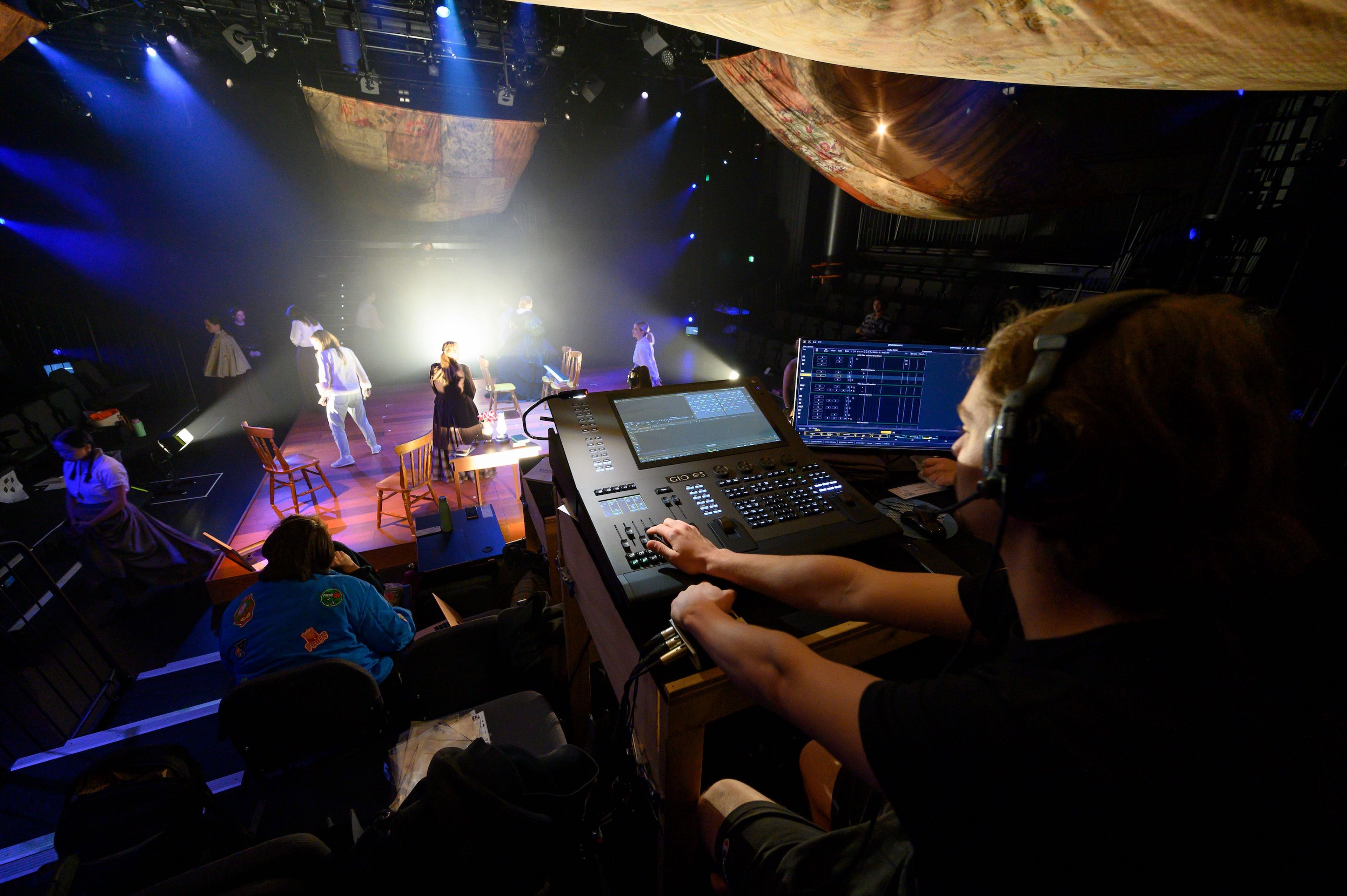 Theatre technician in the control booth operates a lighting console, overlooking a stage filled with actors performing under a warm spotlight.