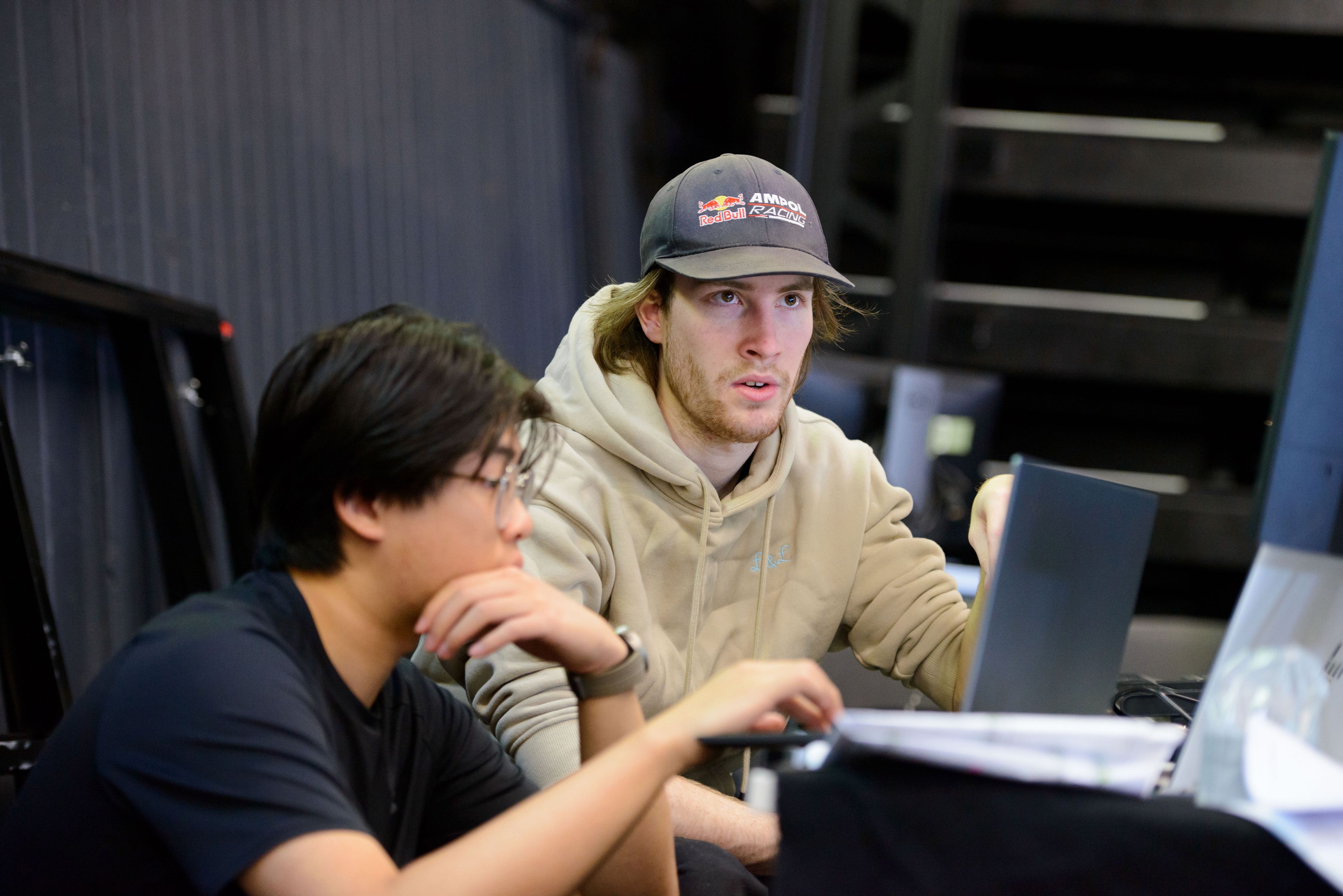 Two individuals focused on a task at laptop, with one wearing a beige hoodie and a cap, while the other, in glasses and a black shirt