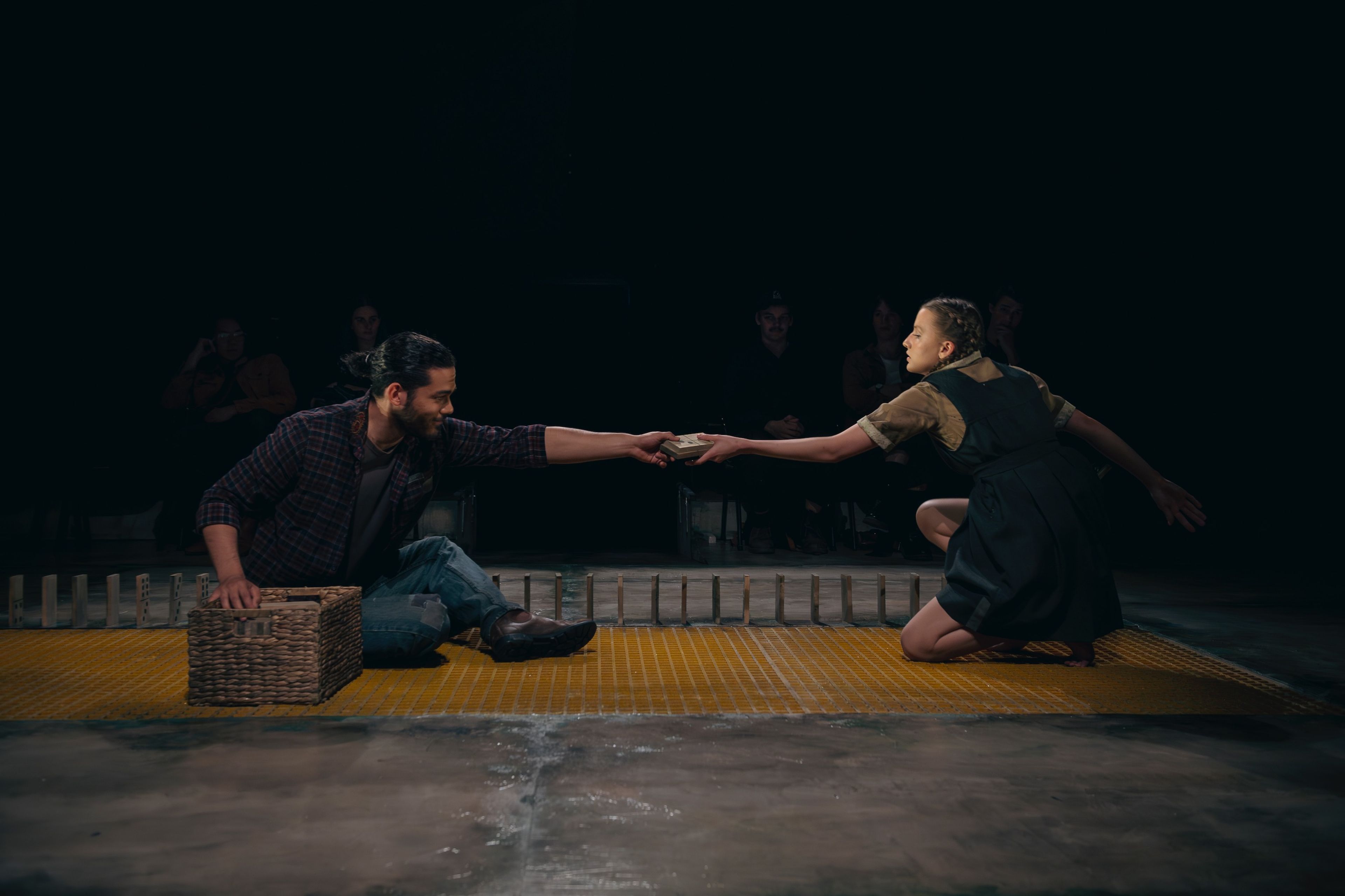 Two performers on a yellow mat, passing an object between them as they stretch towards each other, with one seated beside a wicker basket and the other kneeling.