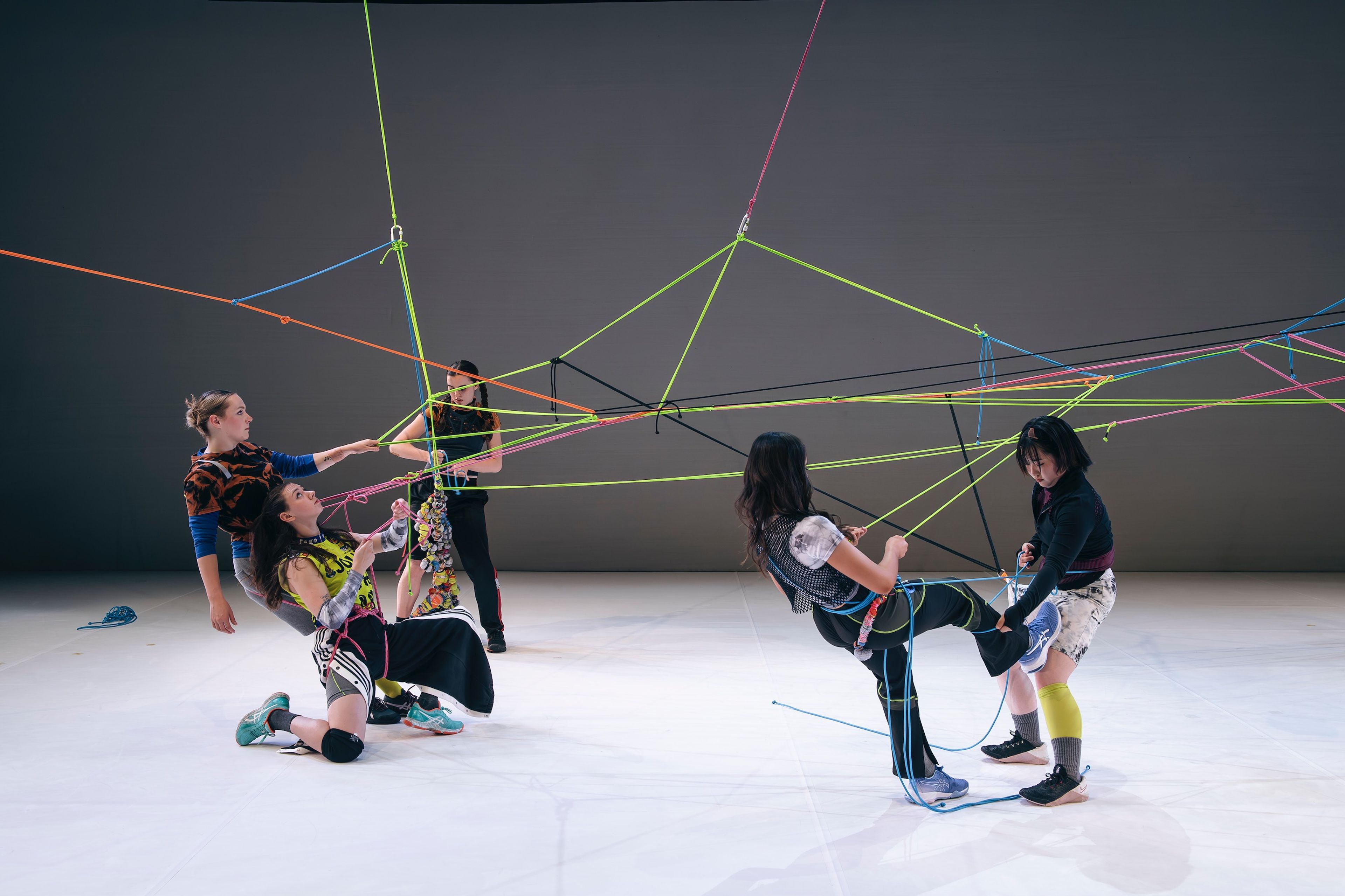 Dancers manipulating a complex network of coloured ropes suspended from above, creating geometric patterns while in various crouching and standing positions on a white floor.