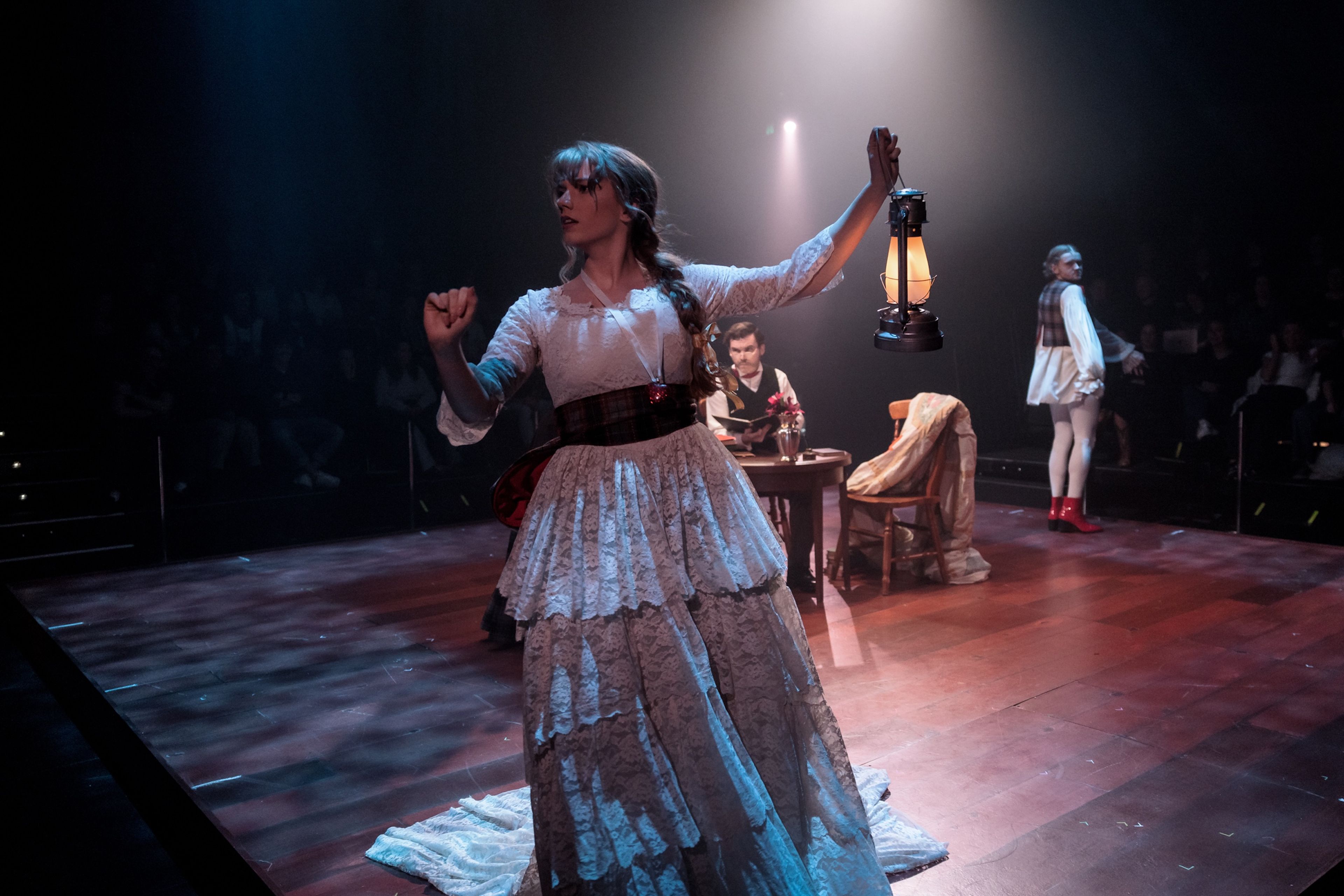 Theatre production photograph featuring a performer in a layered lace dress holding a lantern, with other characters and set pieces visible on a dimly lit stage in the background.