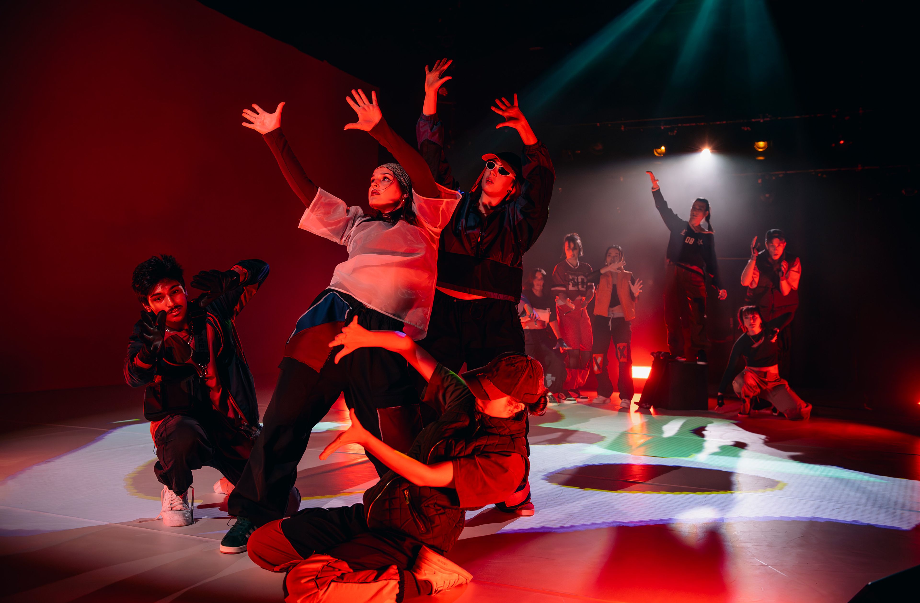 Dance production photograph featuring dancers in energetic poses, with arms raised, bathed in intense red lighting that creates a dramatic atmosphere.