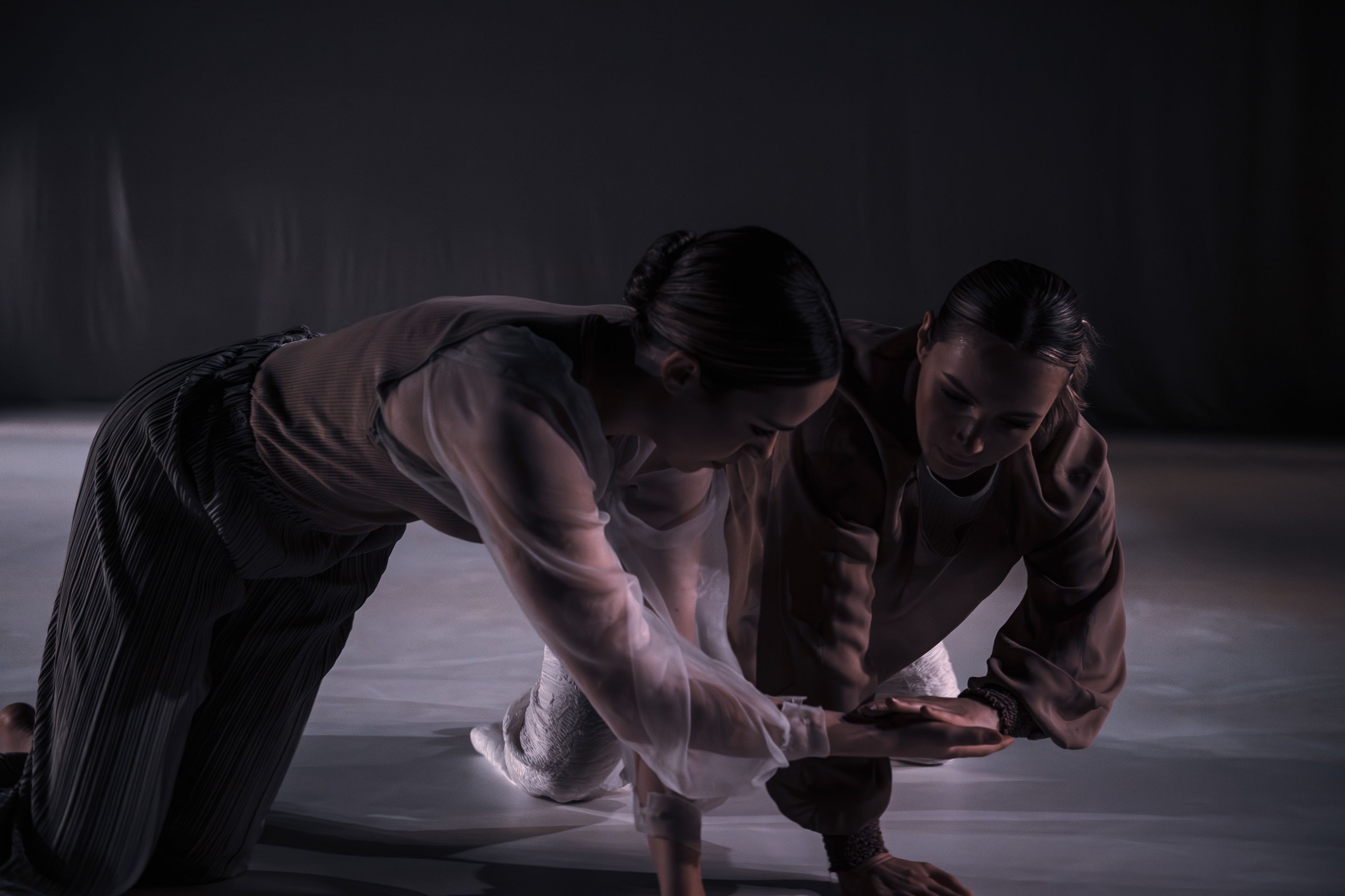 Close-up dance production photograph of two dancers kneeling, their heads bowed together, and soft lighting highlighting their features.