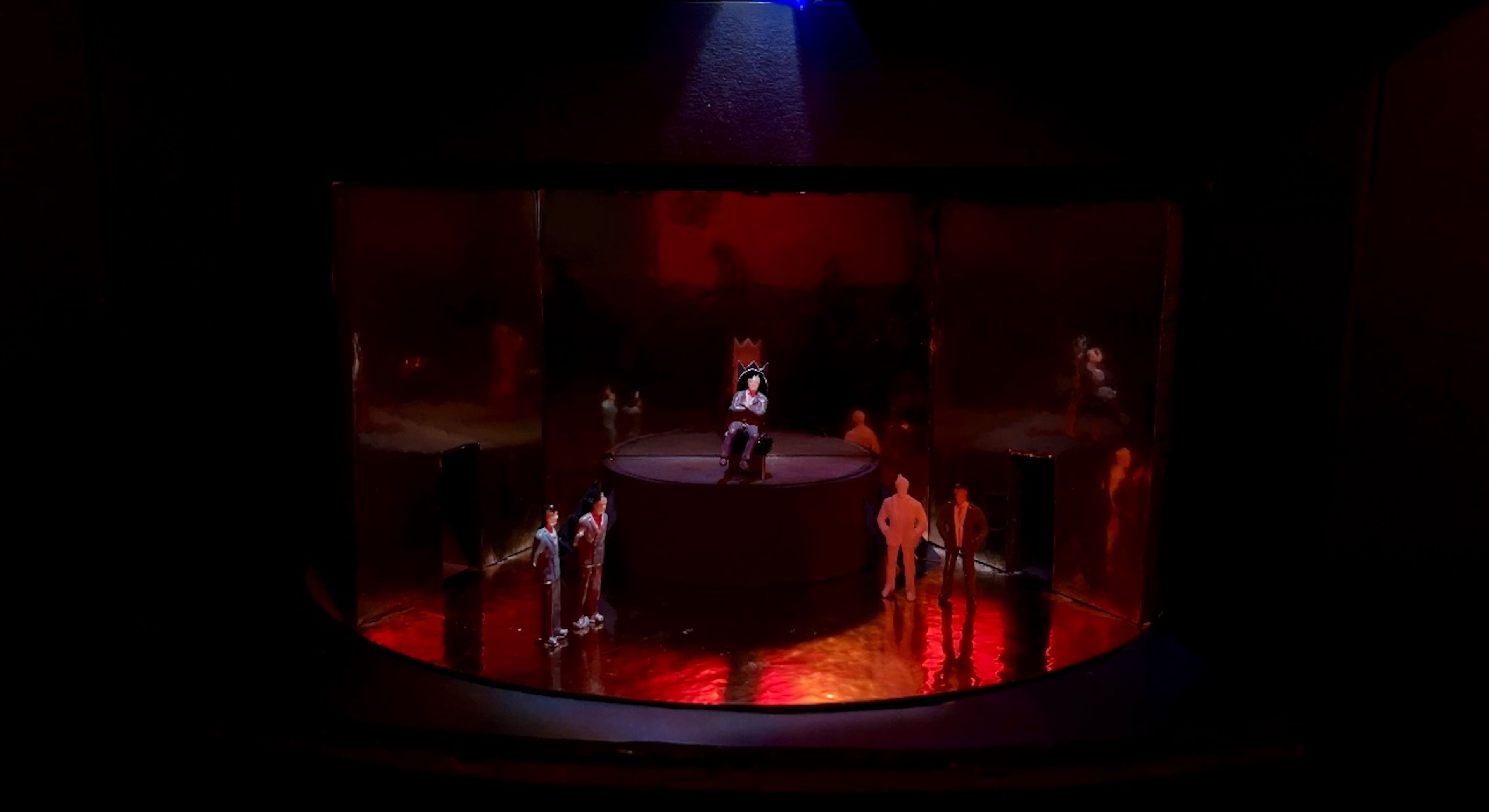 Scale model showing a central figure seated on an elevated platform surrounded by reflective walls, under dim lighting, suggesting isolation and contemplation.