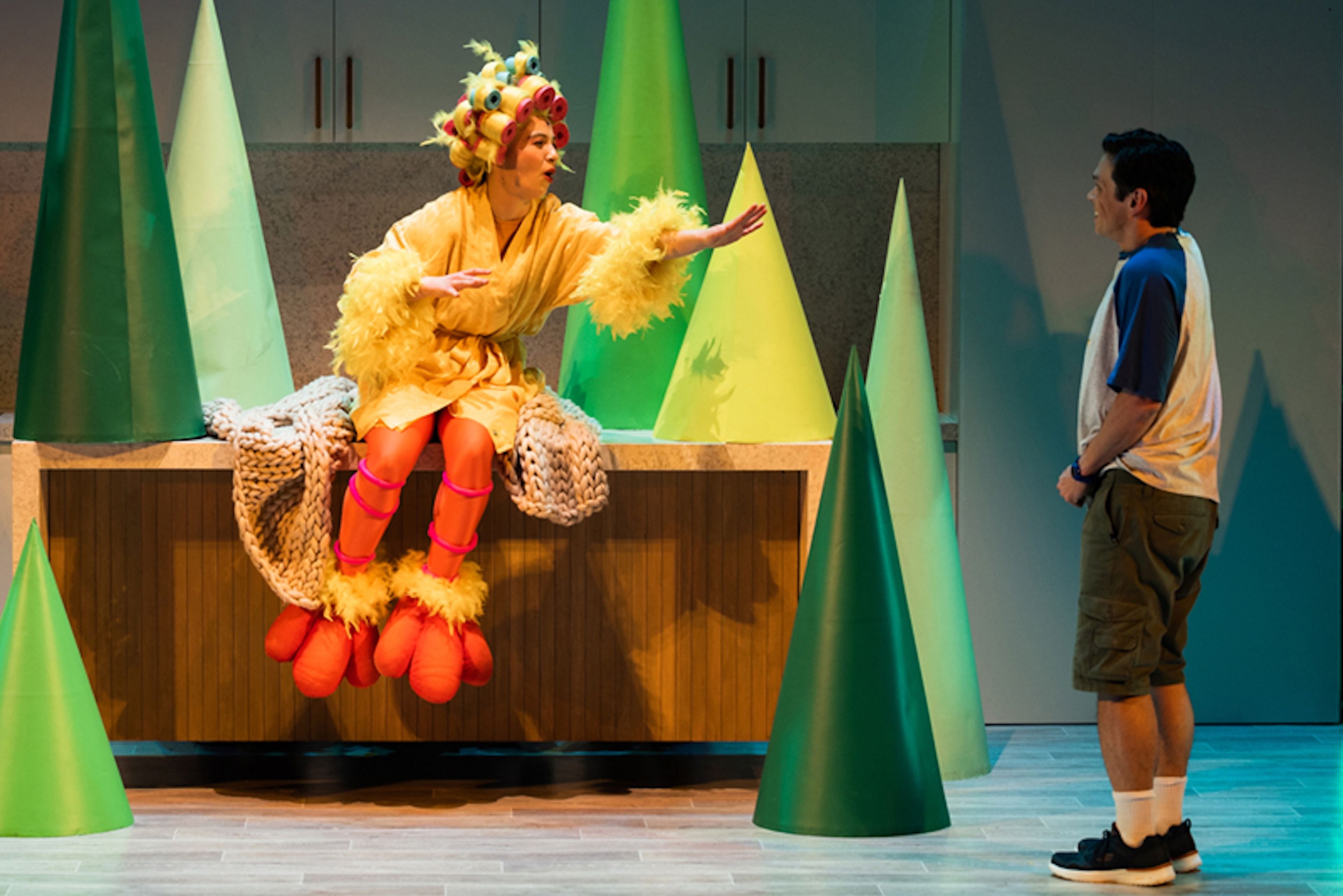 Production photograph of a theatrical scene with a performer in a yellow bird costume with orange legs and colourful hair accessories, seated on a bench among green geometric trees