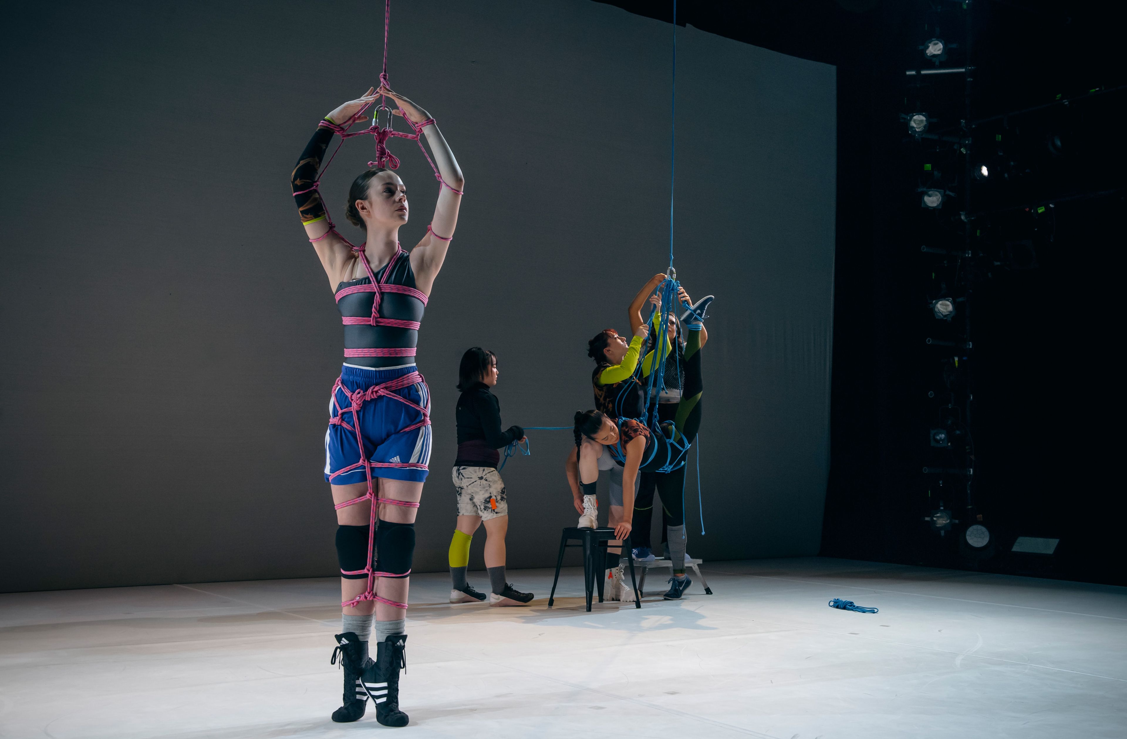 Dance production photograph featuring a dancer suspended in a pink harness, surrounded by other performers adjusting ropes, set against a simple grey backdrop.