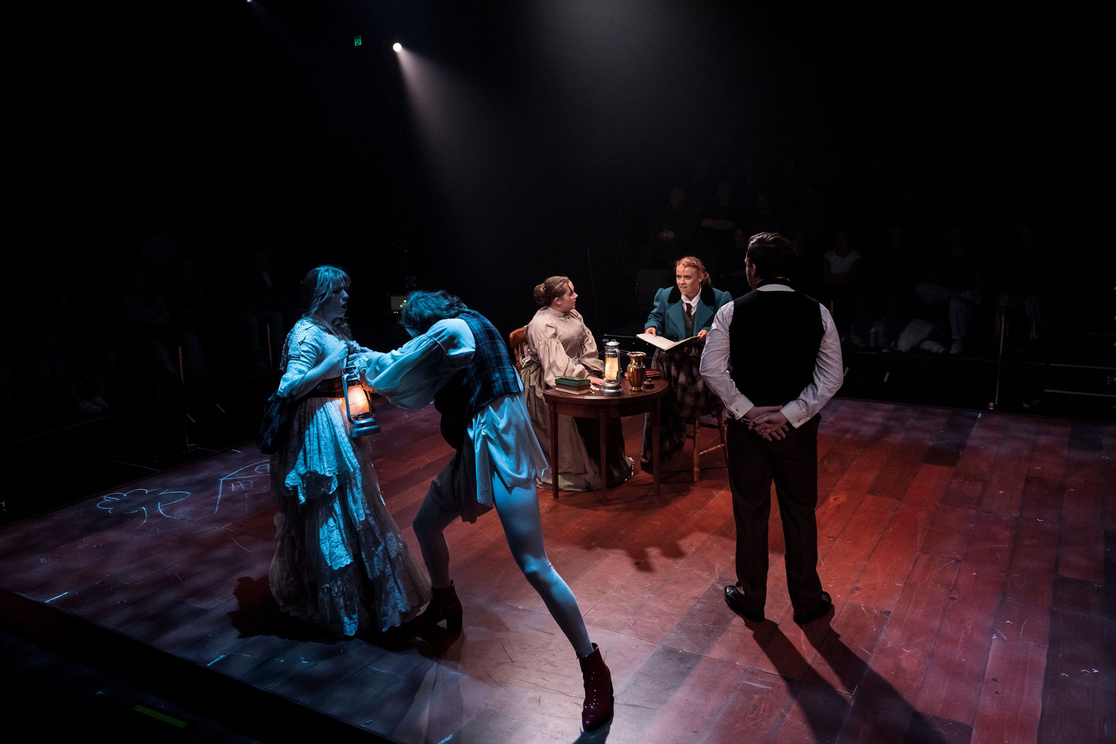 Actors in period costumes sit around a table on a dimly lit stage, with two performers engaged in a dramatic movement nearby.