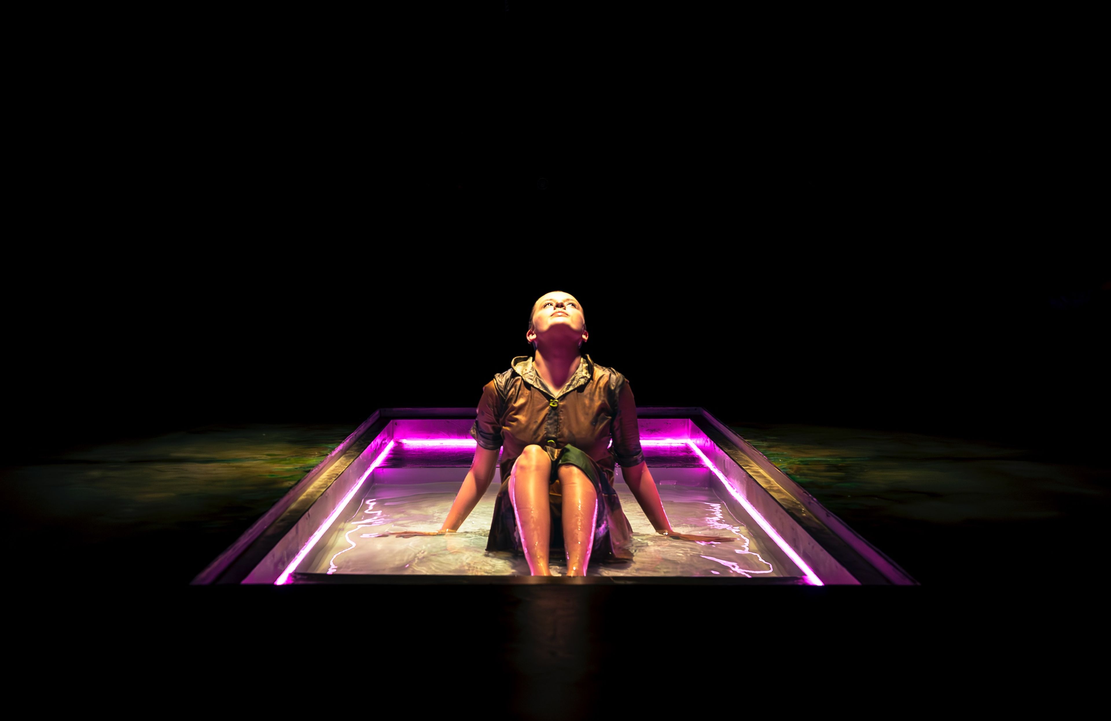 Production photograph of a performer sitting in an illuminated water tank with pink LED lighting, looking upward against a black background