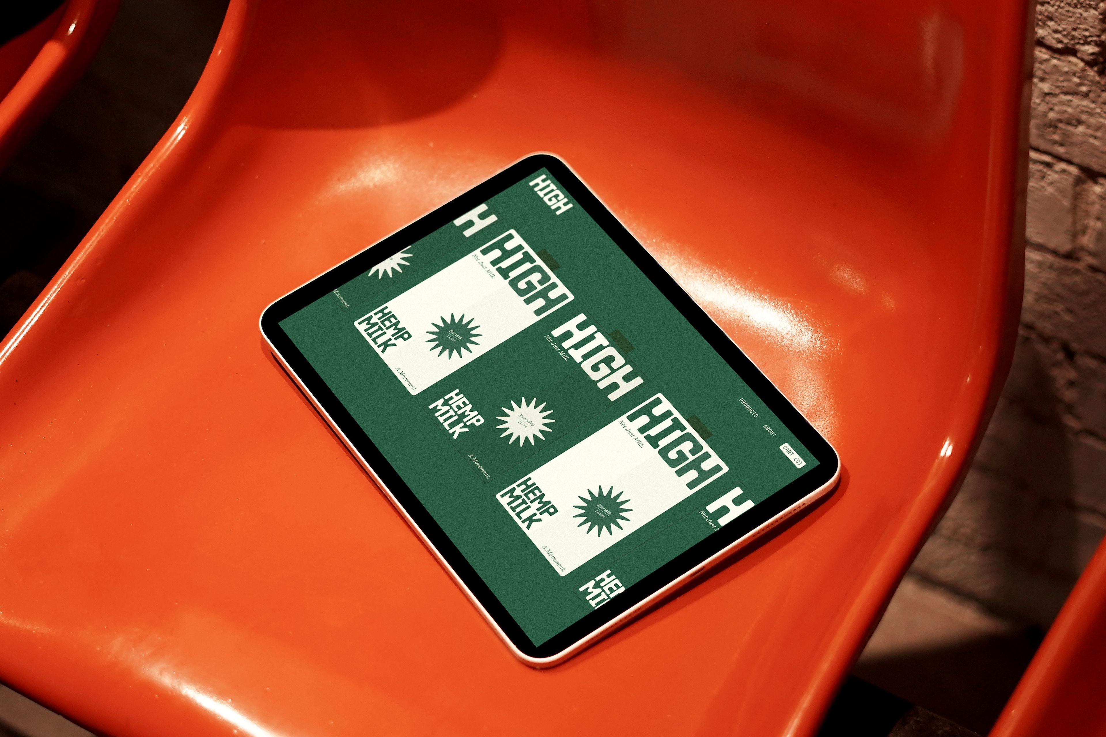 Digital mockup of 'Hemp Milk' branding displayed on a tablet, resting on an orange plastic chair. The green and white design features bold typography with the word 'HIGH' and 'HEMP MILK' alongside a stylised sunburst graphic.