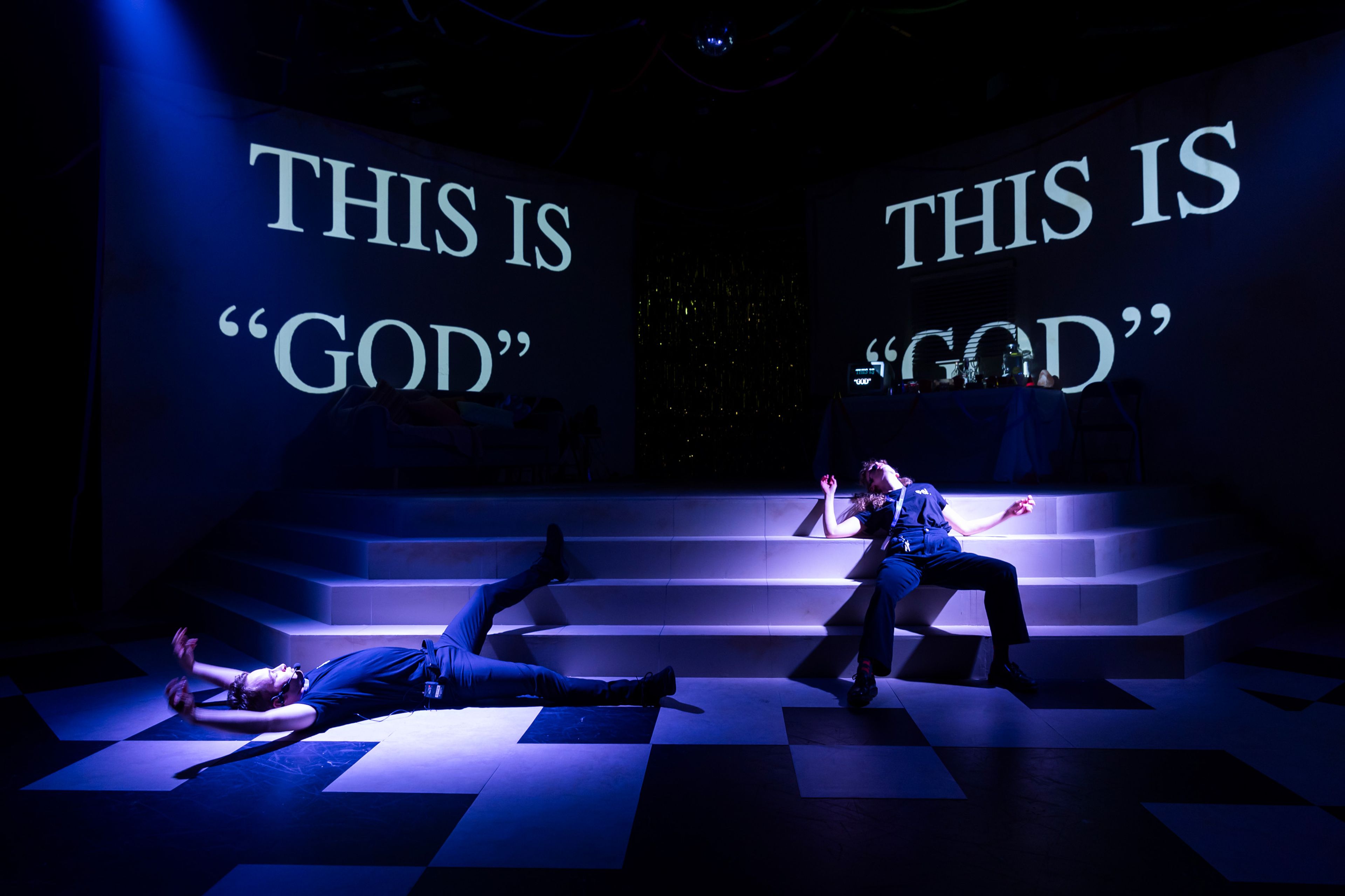 Theatre production photograph showing two performers arranged on a black and white checkerboard floor with stepped platforms, beneath projected text reading 'THIS IS "GOD"' lit in cool blue tones.