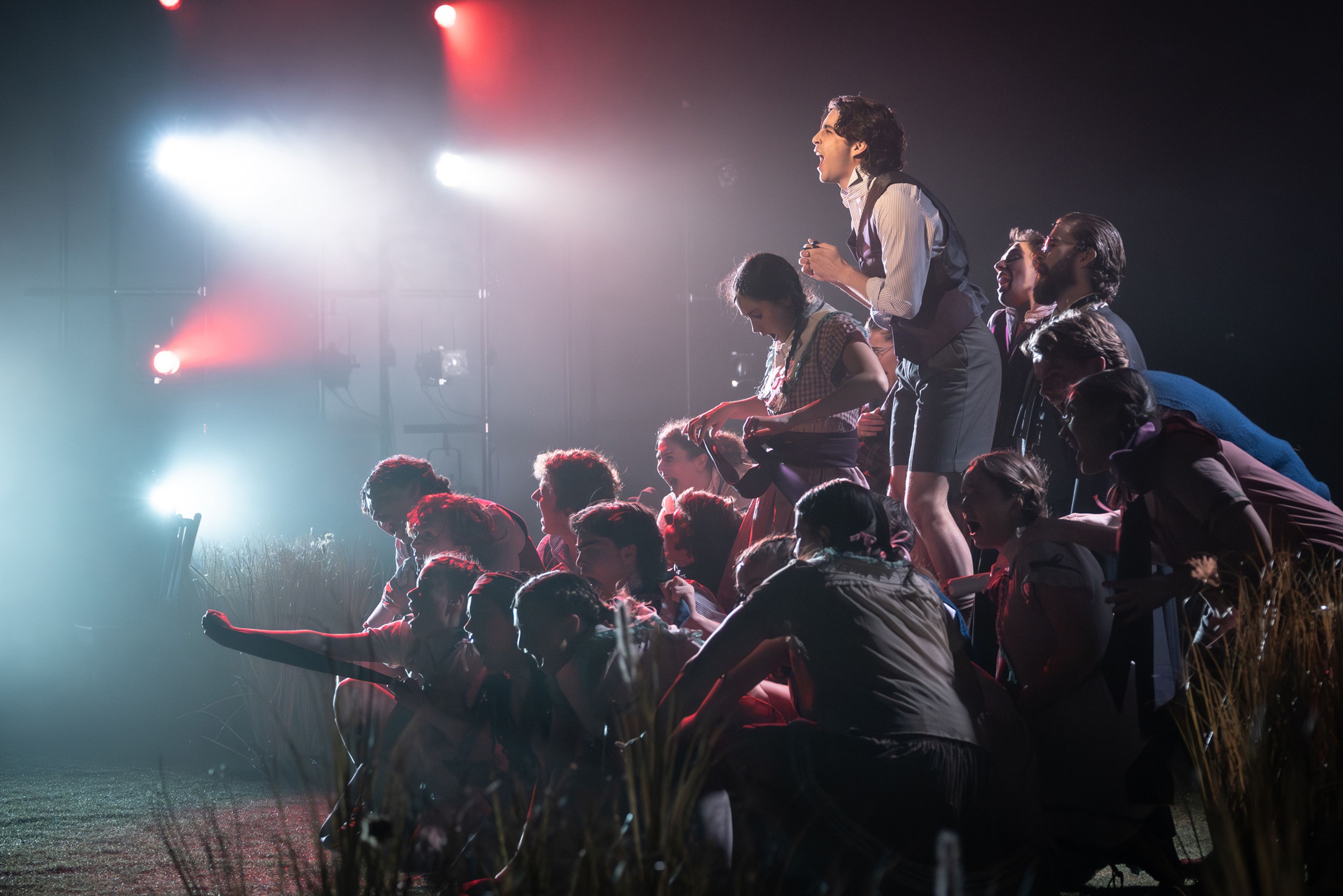 Theatre production photograph showing a dramatic ensemble moment with performers arranged in ascending levels, with one highlighted performer standing above the group, lit by dramatic white and red theatrical lighting through haze.