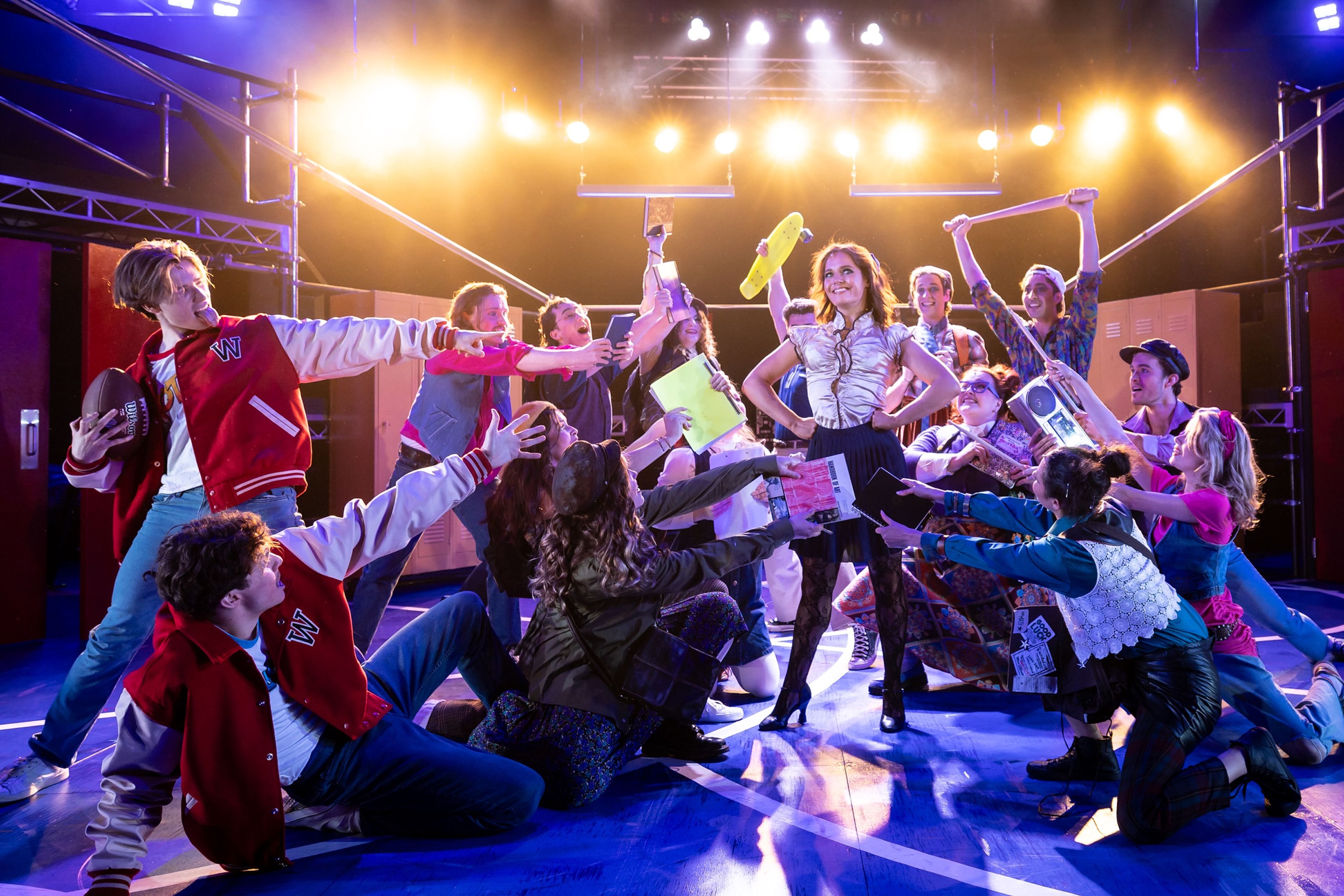 A lively dance scene set in a high school environment. Characters in '80s-themed costumes, including letterman jackets and cheerleader outfits, gather around a central female figure.