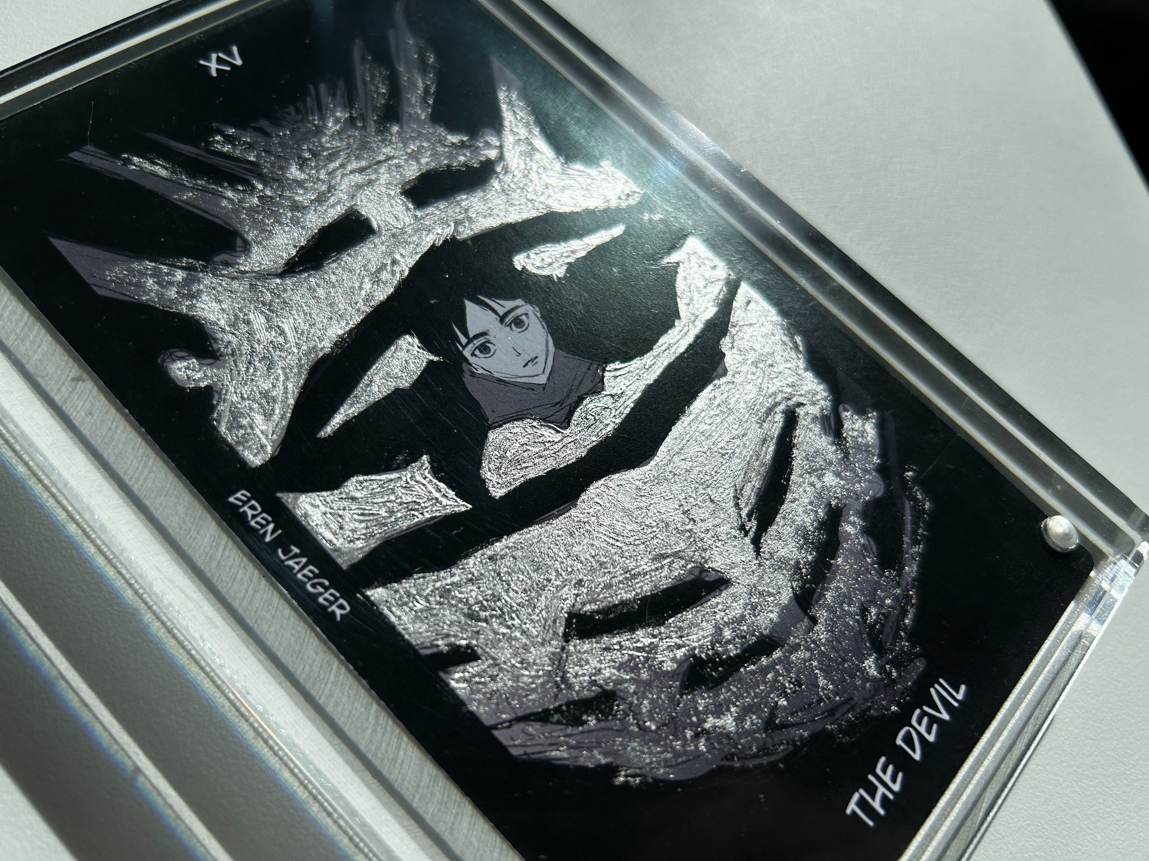 Close-up of a tarot card titled "The Devil" featuring a sombre portrait of a character named Eren Jaeger, with a textured silver overlay surrounding the figure, adding a dark, metallic effect to the design.