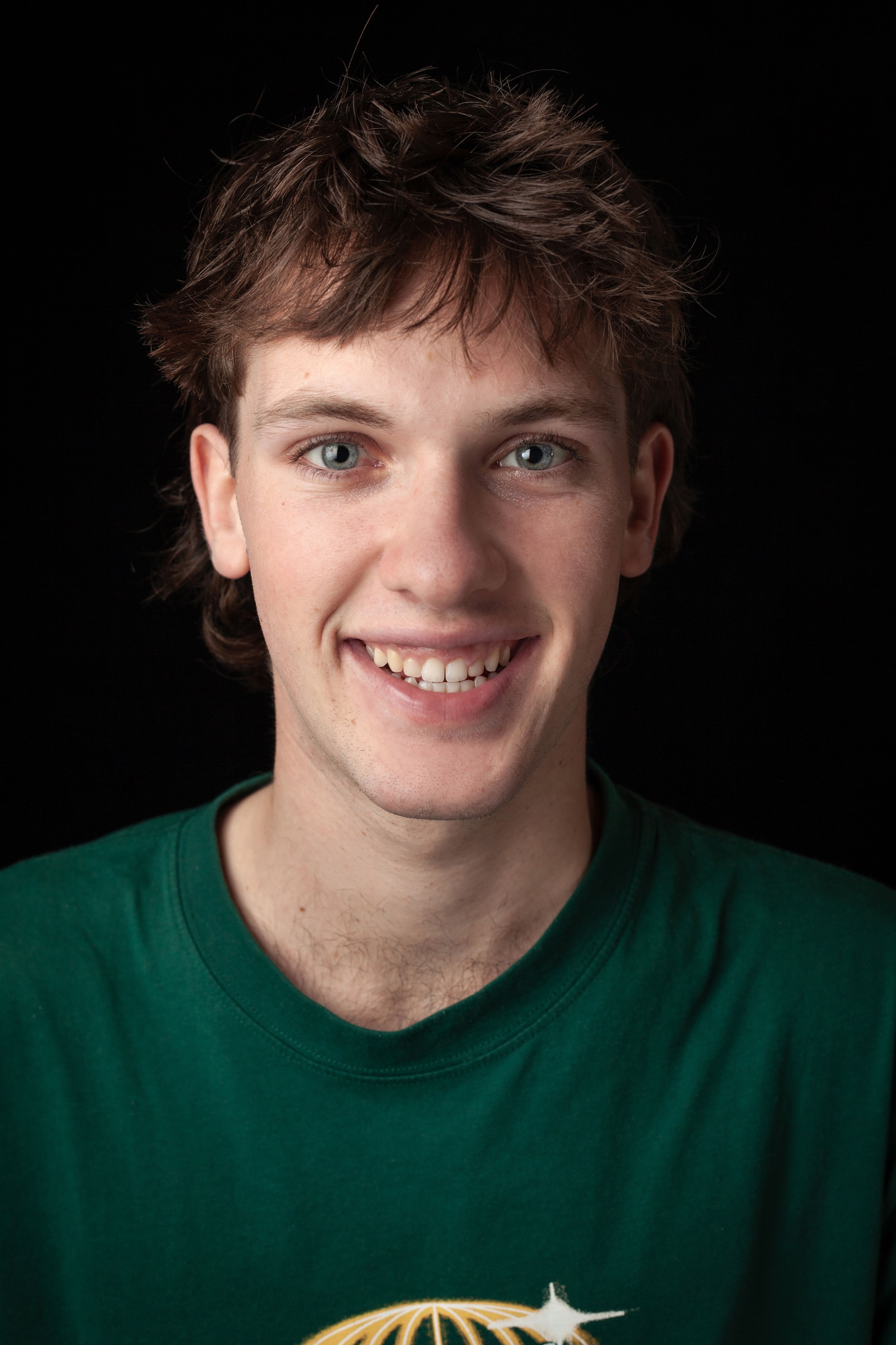 Headshot of a smiling young man with shaggy brown hair, wearing a green T-shirt, against a black background.