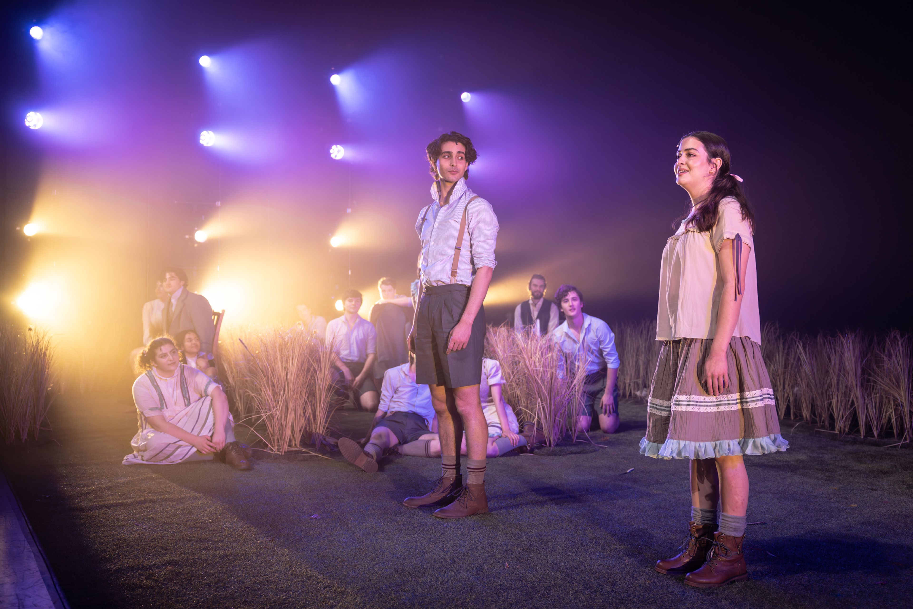 Theatre production photograph showing performers in period costumes in a dreamlike scene, with dramatic purple and amber lighting creating atmospheric beams above the wheat grass set.