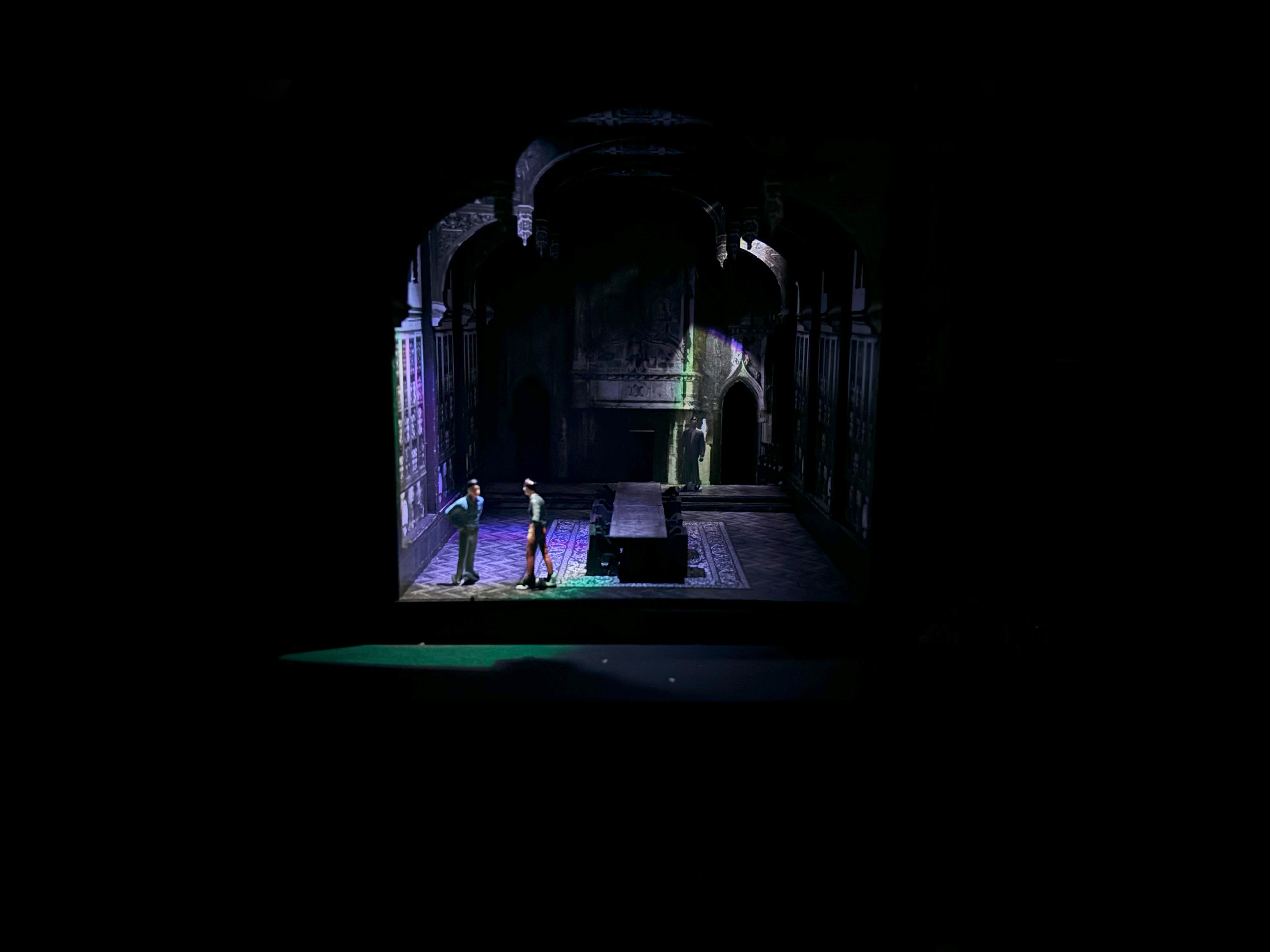 
The image shows a theatrical set model featuring a dimly lit medieval-style hall with high arched architecture and intricate details. A long central table is flanked by two miniature figures engaged in interaction