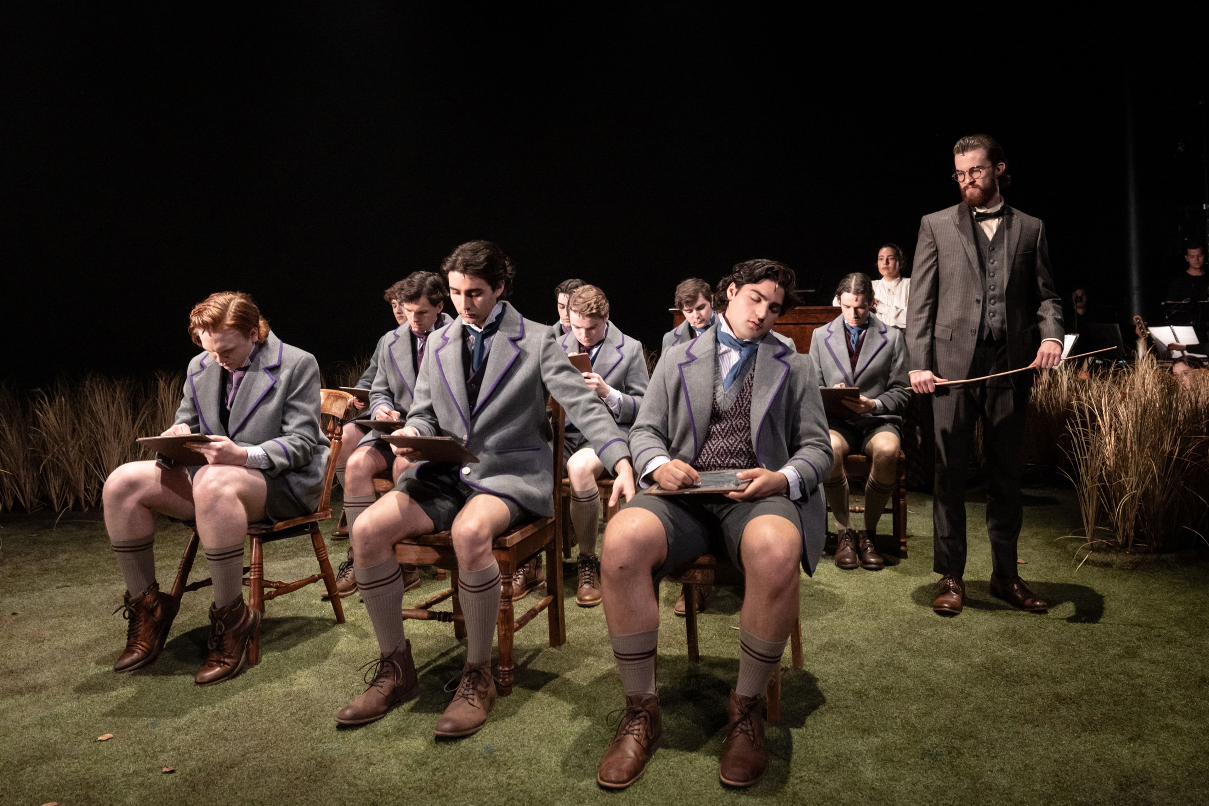 Theatre production photograph showing schoolboys in matching grey uniforms with purple trim seated in wooden chairs, studying or writing, with an adult figure standing at the end of the row and wheat grass visible in the background.