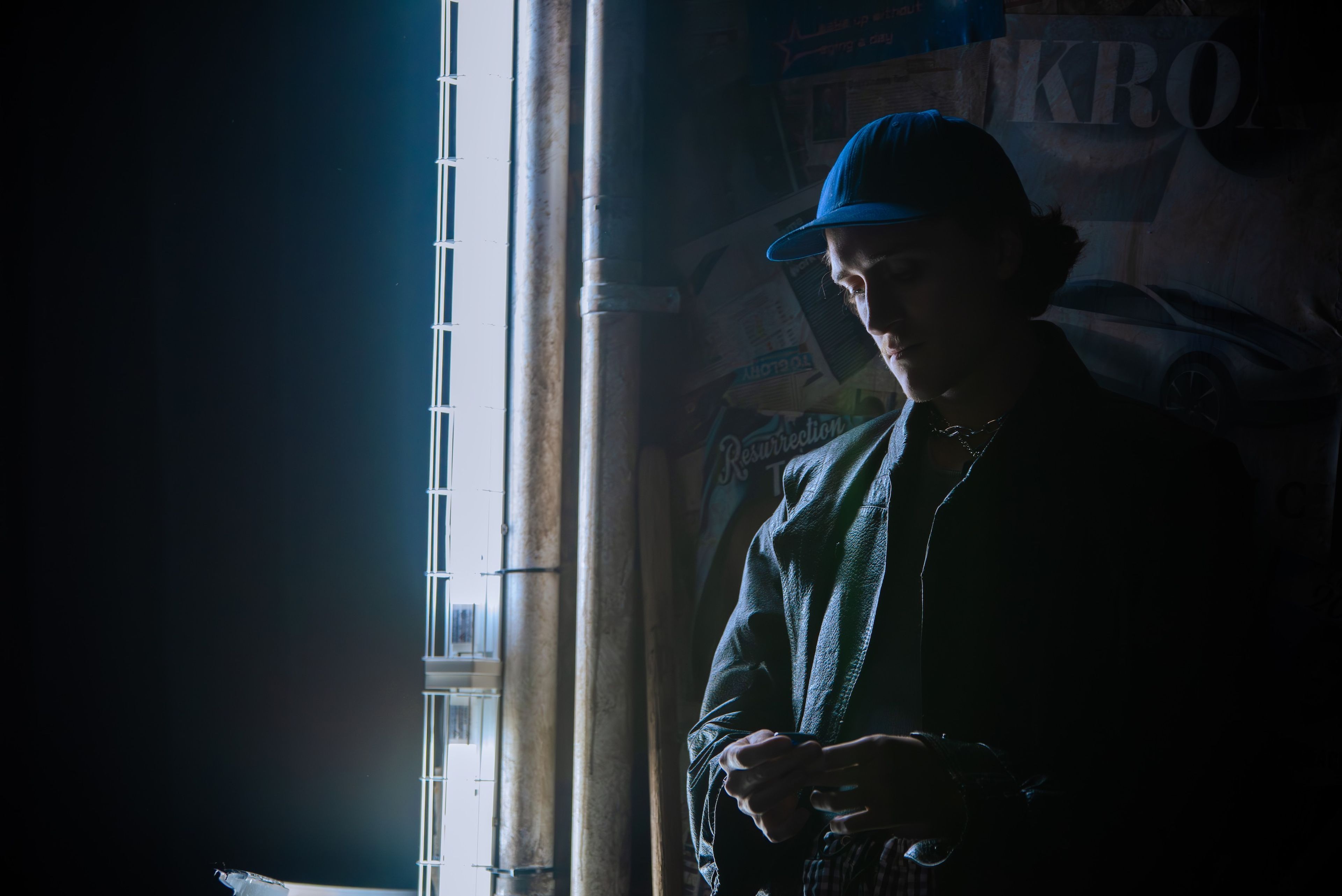 Theatre production photograph featuring a character in a blue cap, standing pensively beside a brightly lit window, casting soft shadows in a dimly lit space.